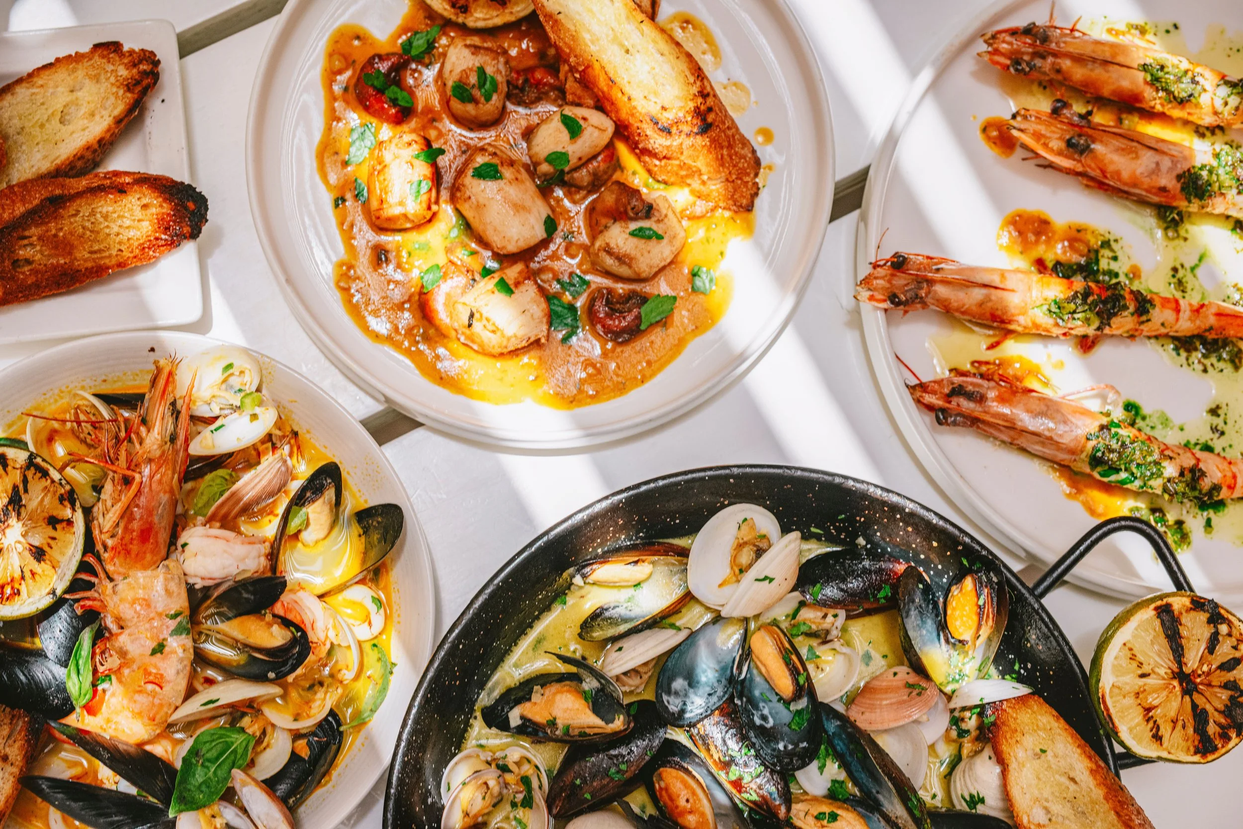 Assorted seafood dishes including shrimp, clams, mussels, and grilled shrimp, served with toasted bread and lemon halves.