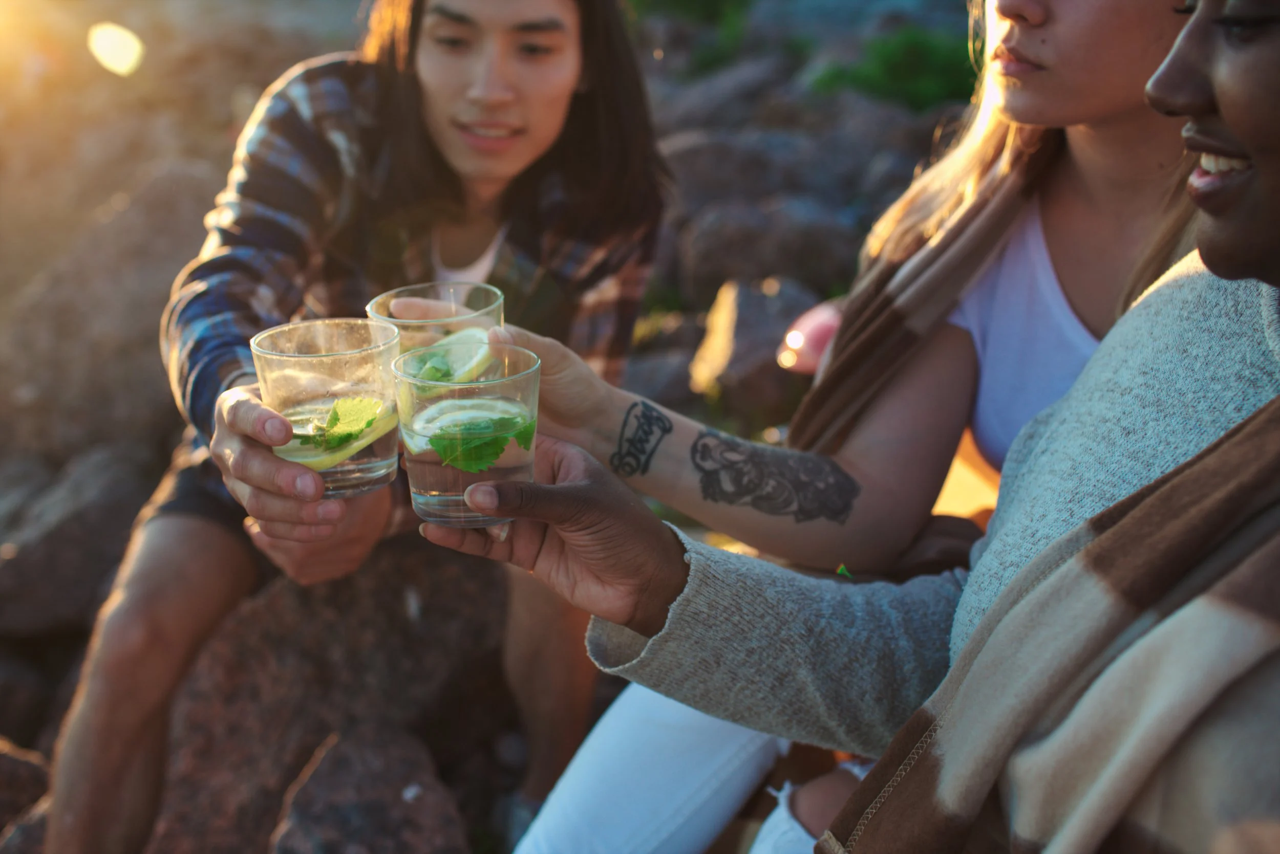 People clinking glasses with drinks containing mint leaves outdoors during sunset.