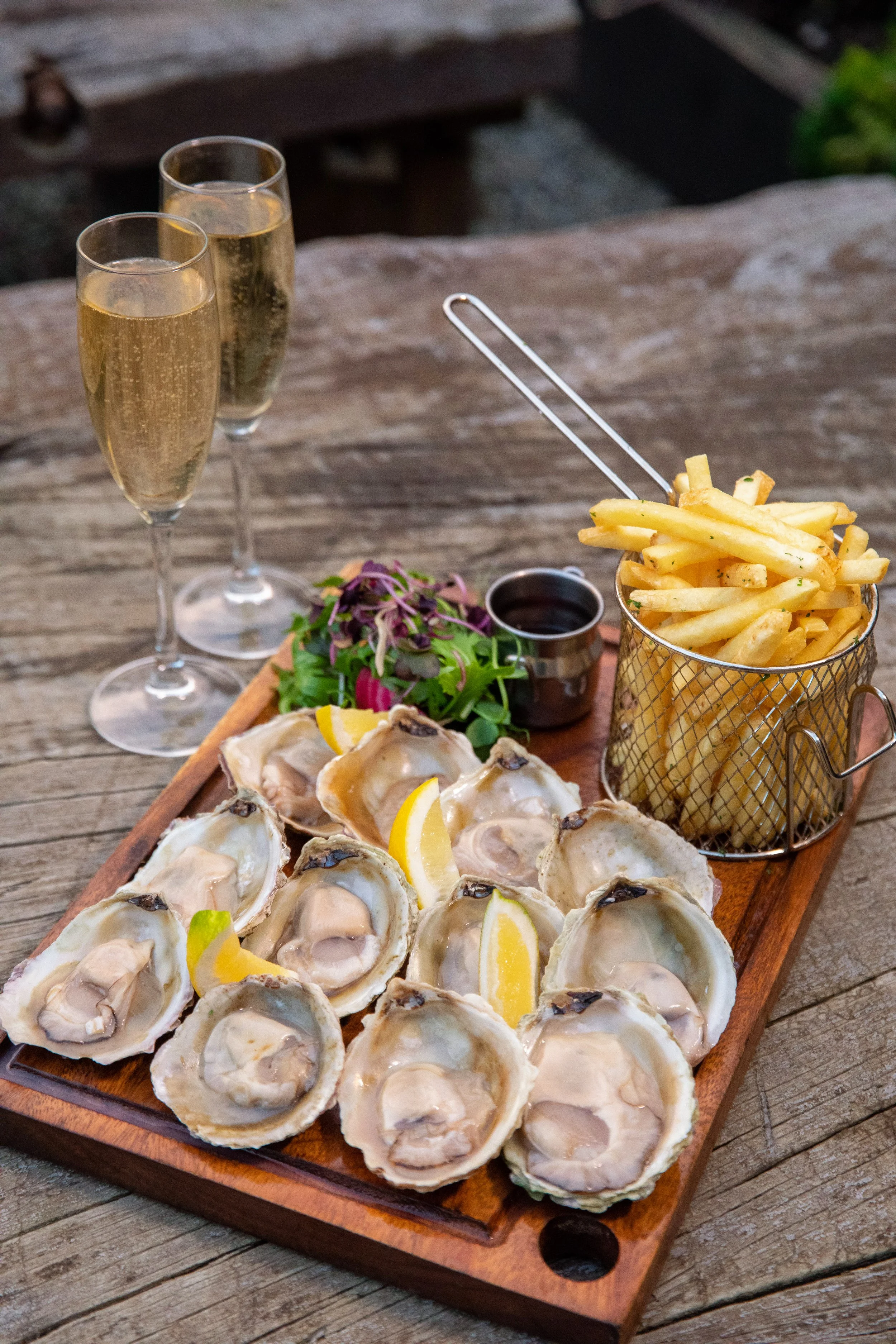 A wooden serving board with raw oysters on the half shell garnished with lemon wedges, a small salad, and a container of French fries, accompanied by two glasses of champagne.