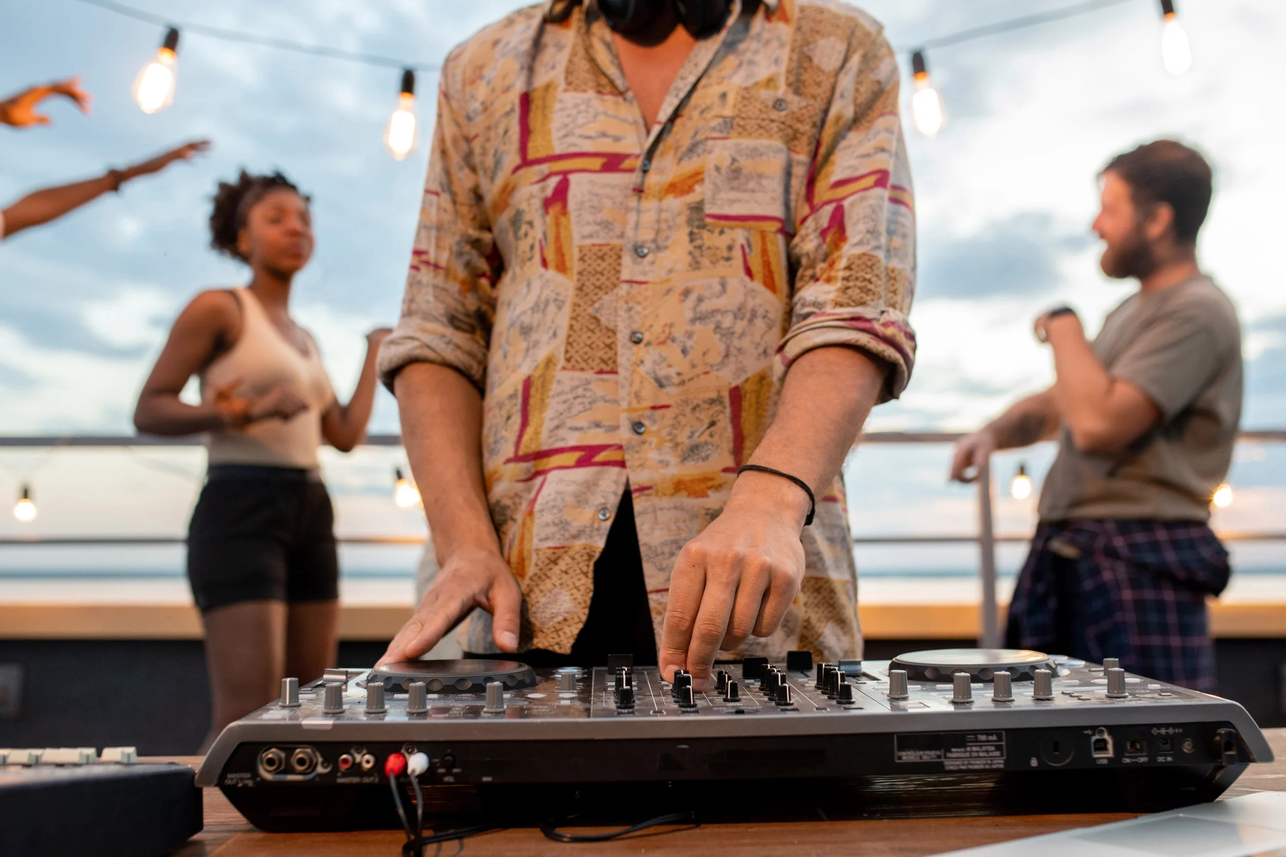 Person in a patterned shirt DJing at an outdoor gathering during sunset with string lights and people dancing in the background.