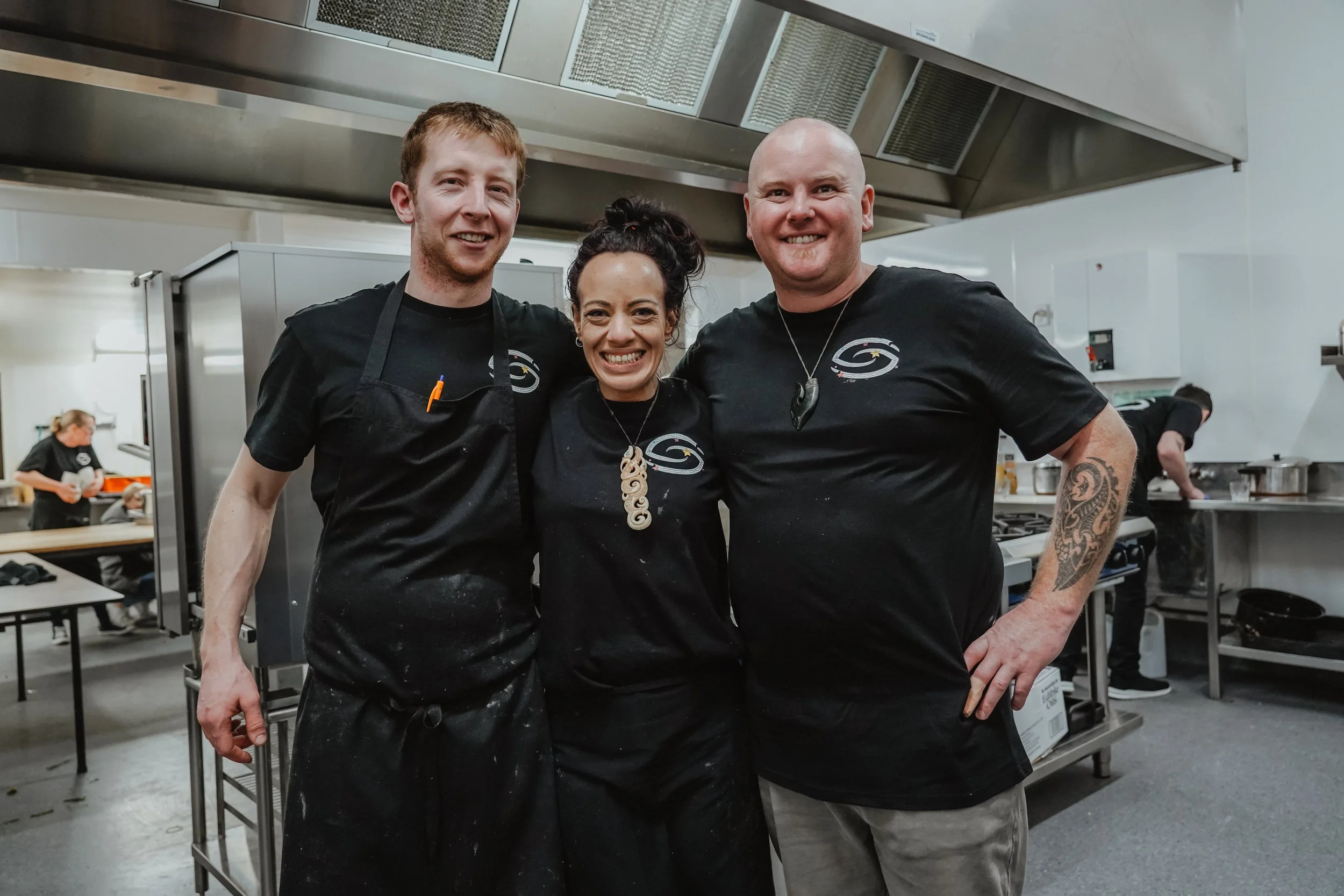 Three people smiling in a commercial kitchen, wearing black shirts with a logo, surrounded by cooking equipment.