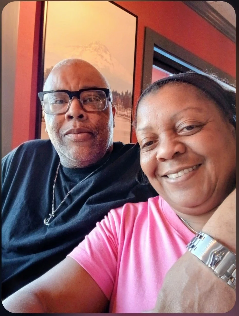 A man and woman taking a close-up selfie inside a restaurant. The man is wearing glasses and a black shirt, and the woman is smiling, wearing a pink shirt and a silver watch. There are framed pictures on the yellowish-orange wall behind them.