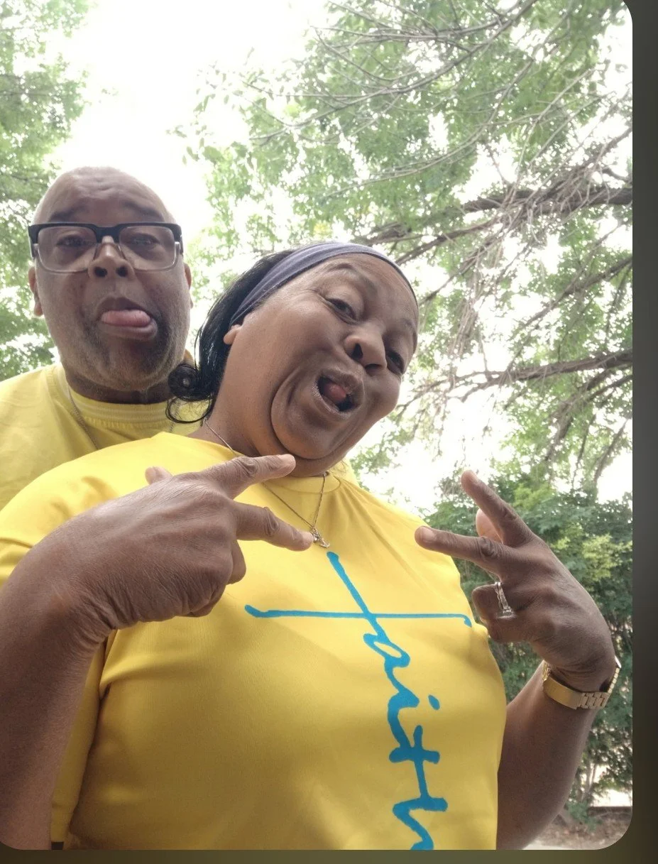 Two adults outdoors under trees, making playful faces and peace signs, wearing matching yellow shirts, with a background of green leaves.