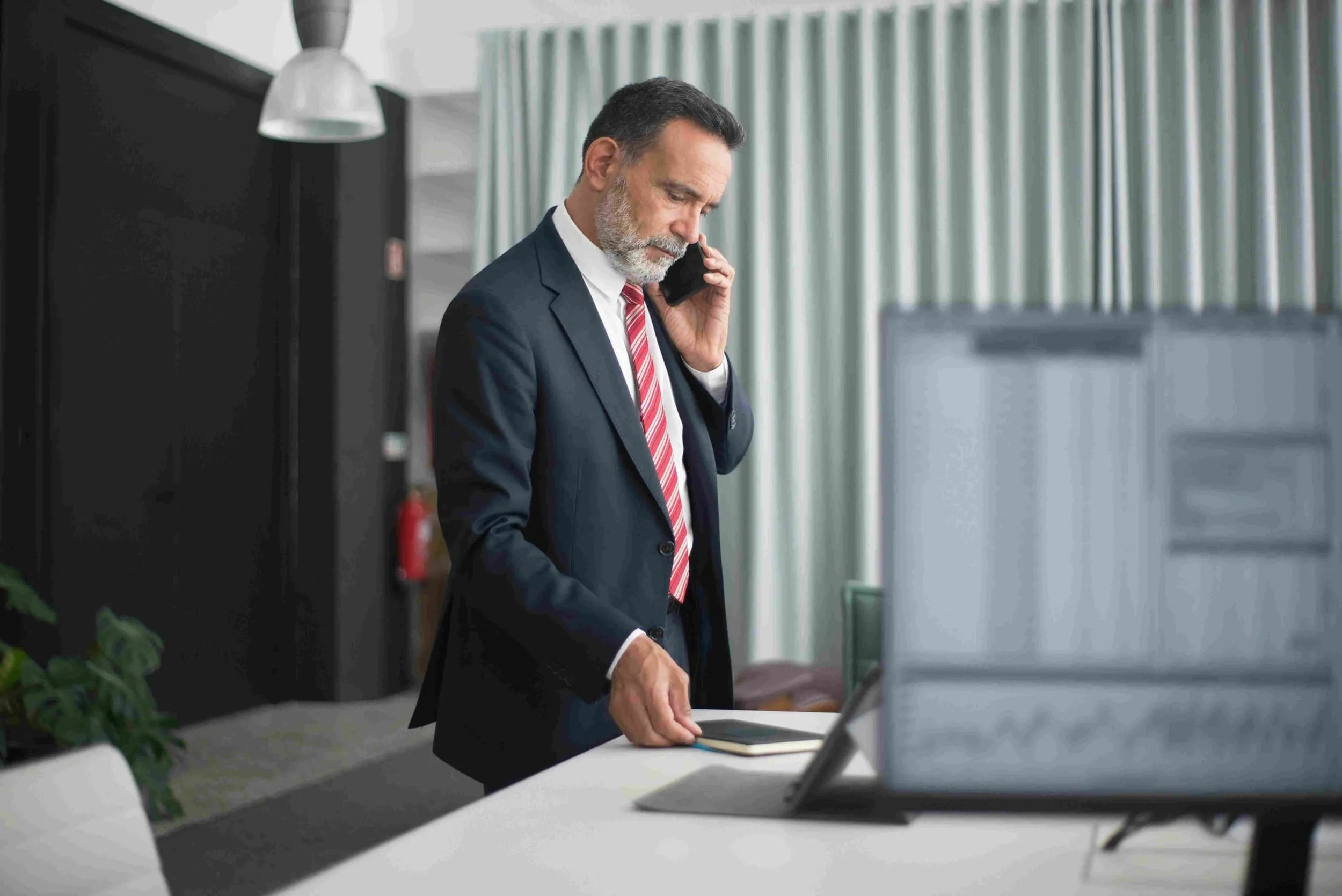 Regenerate alt text copyIconcrossIconBlue Businessman in a suit discusses hiring on the phone while standing at his desk with a laptop and documents in a modern office.