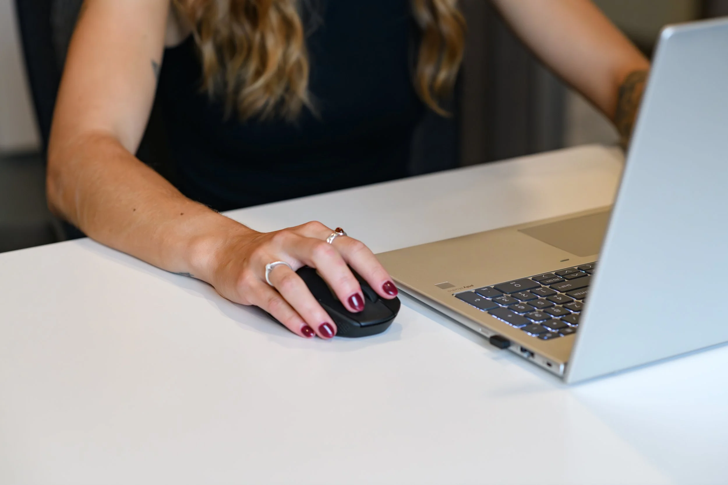 A woman with long, wavy blonde hair working at a white desk, using a black computer mouse and a silver laptop.