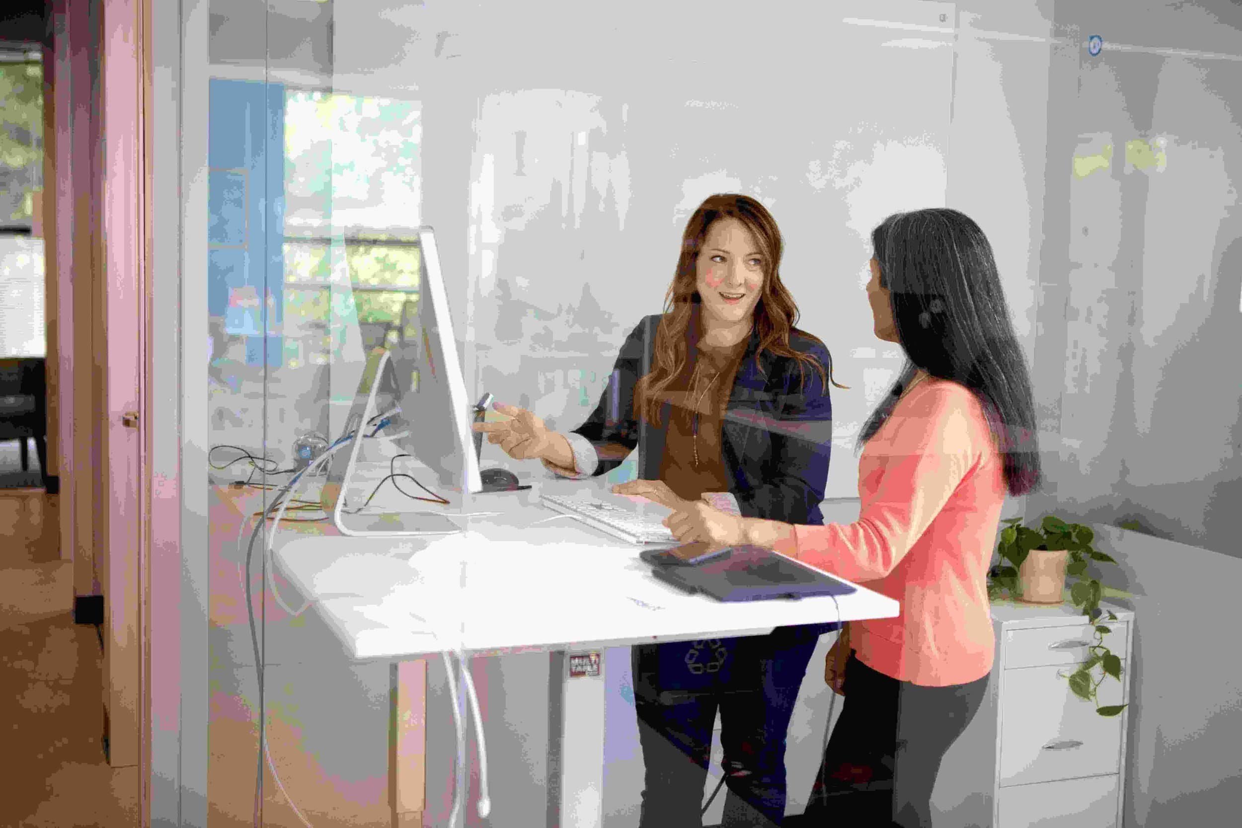 Two women standing and smiling, collaborating with a recruiter at a computer in a bright, modern office.