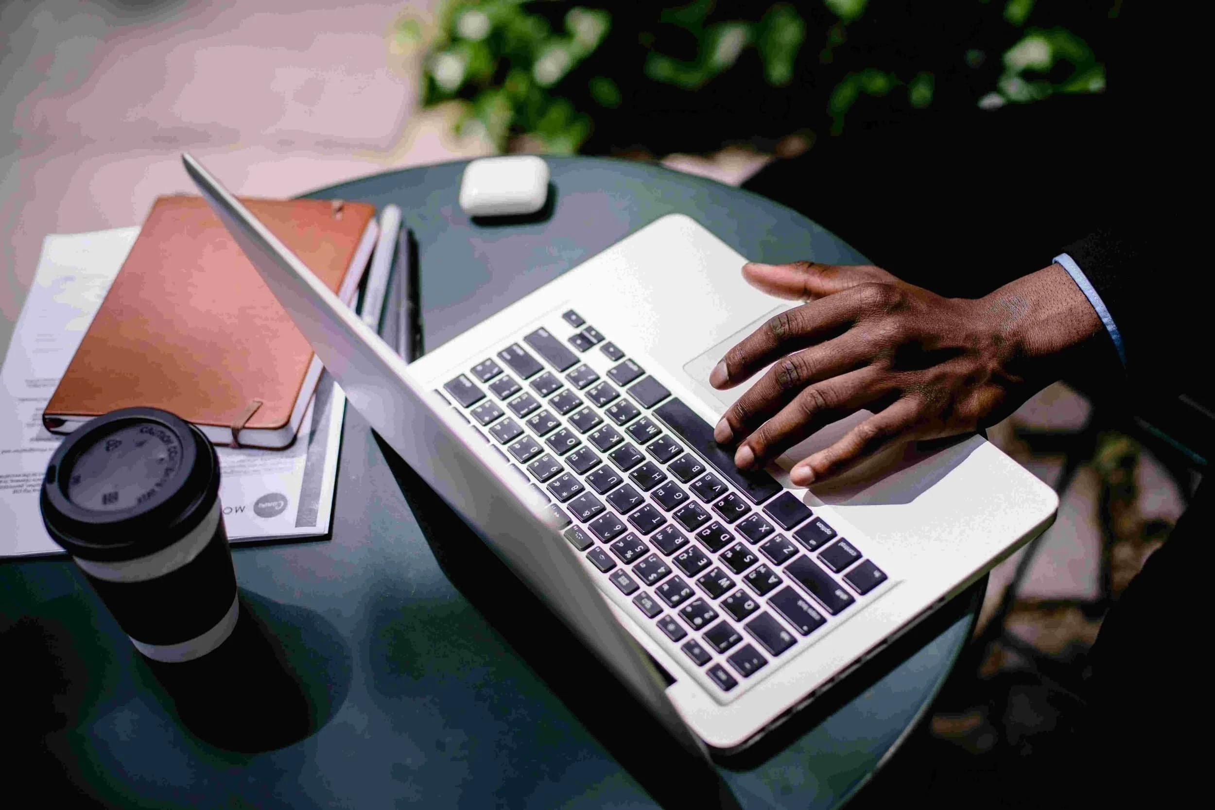 A person recruiting on a laptop at an outdoor table with a notebook, documents, coffee cup, and earbuds nearby.