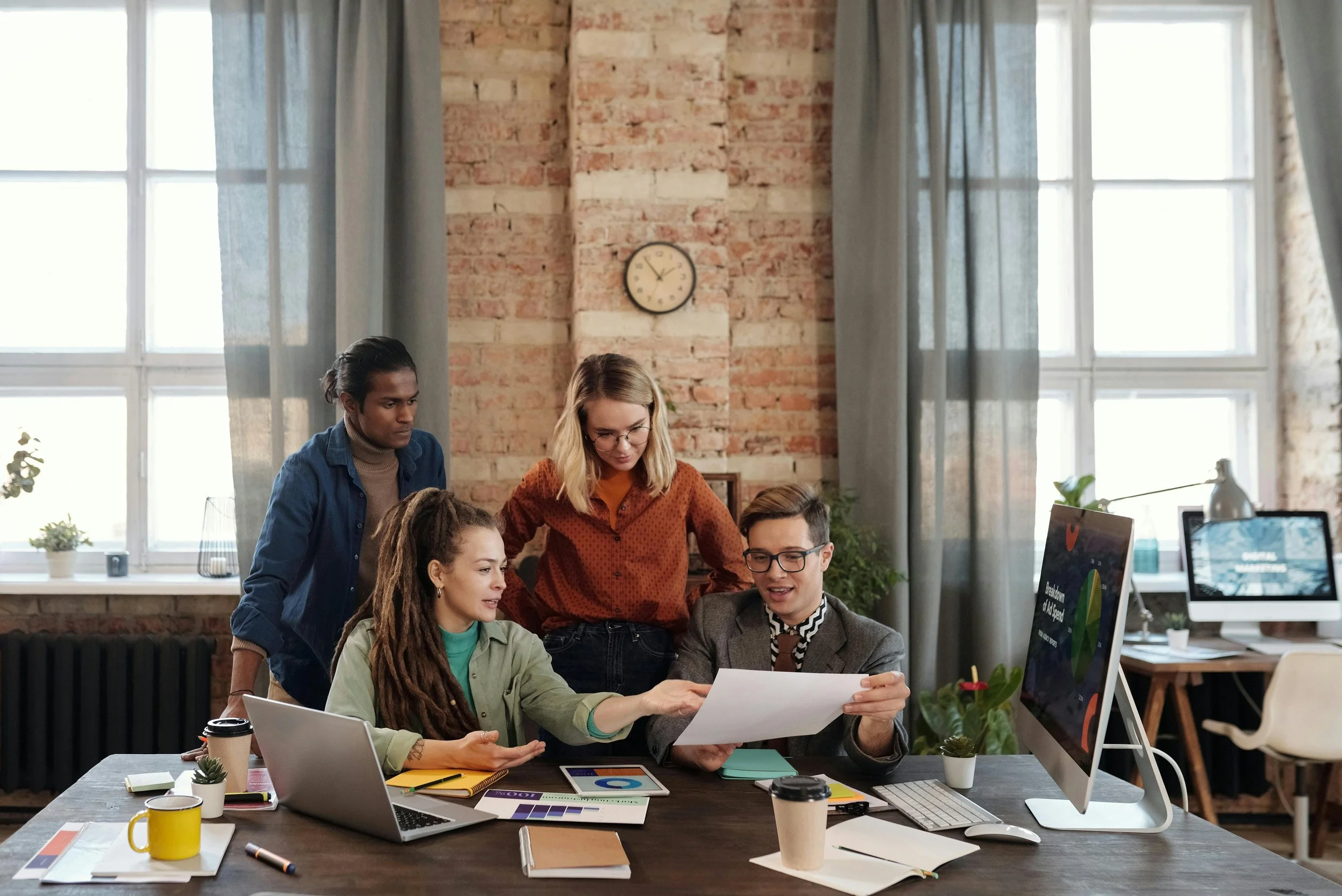 Four young professionals collaborating around a desk in a modern office with large windows, a brick wall, and various office supplies and electronic devices.