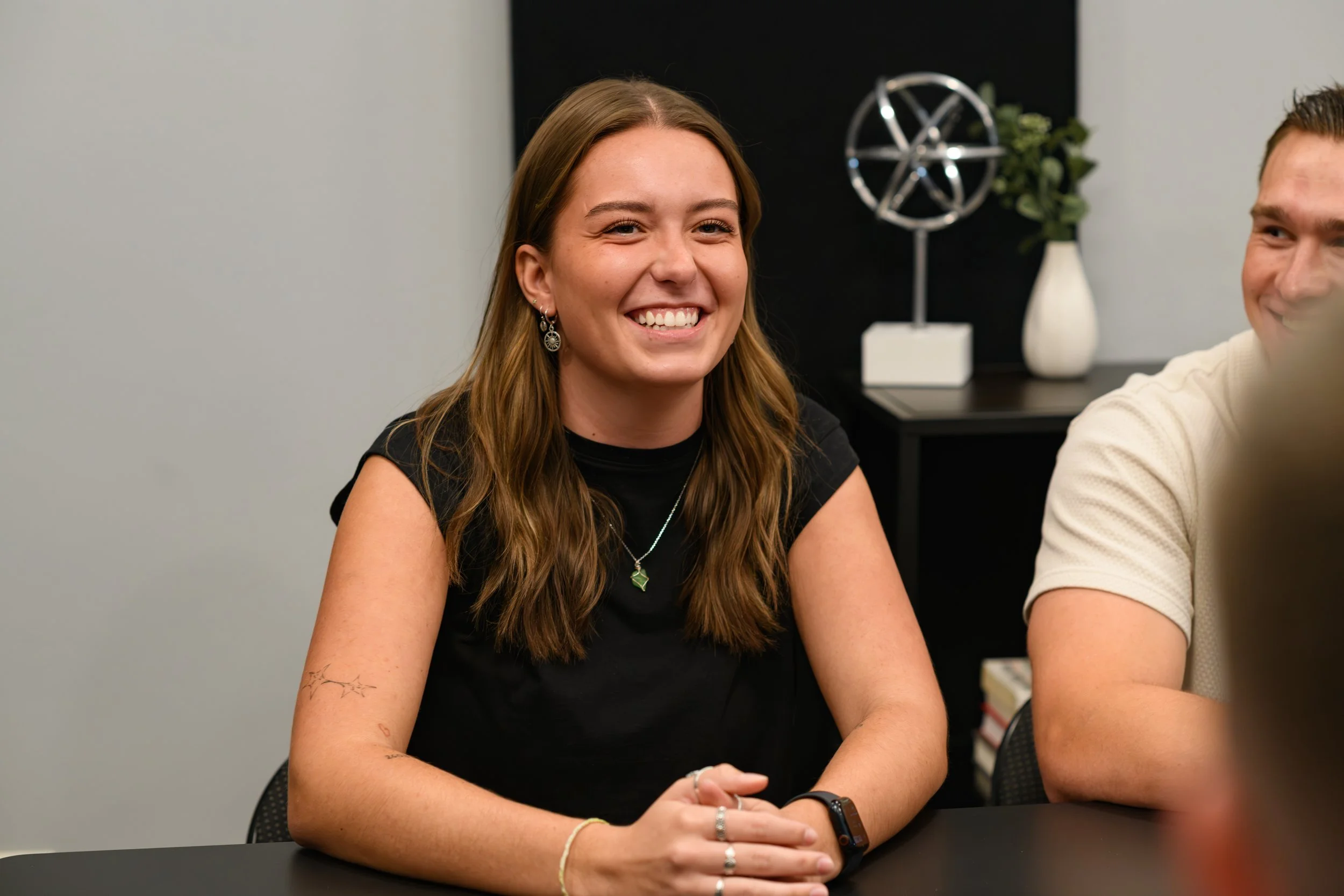 A young woman with long brown hair smiling, sitting at a table in an indoor setting.