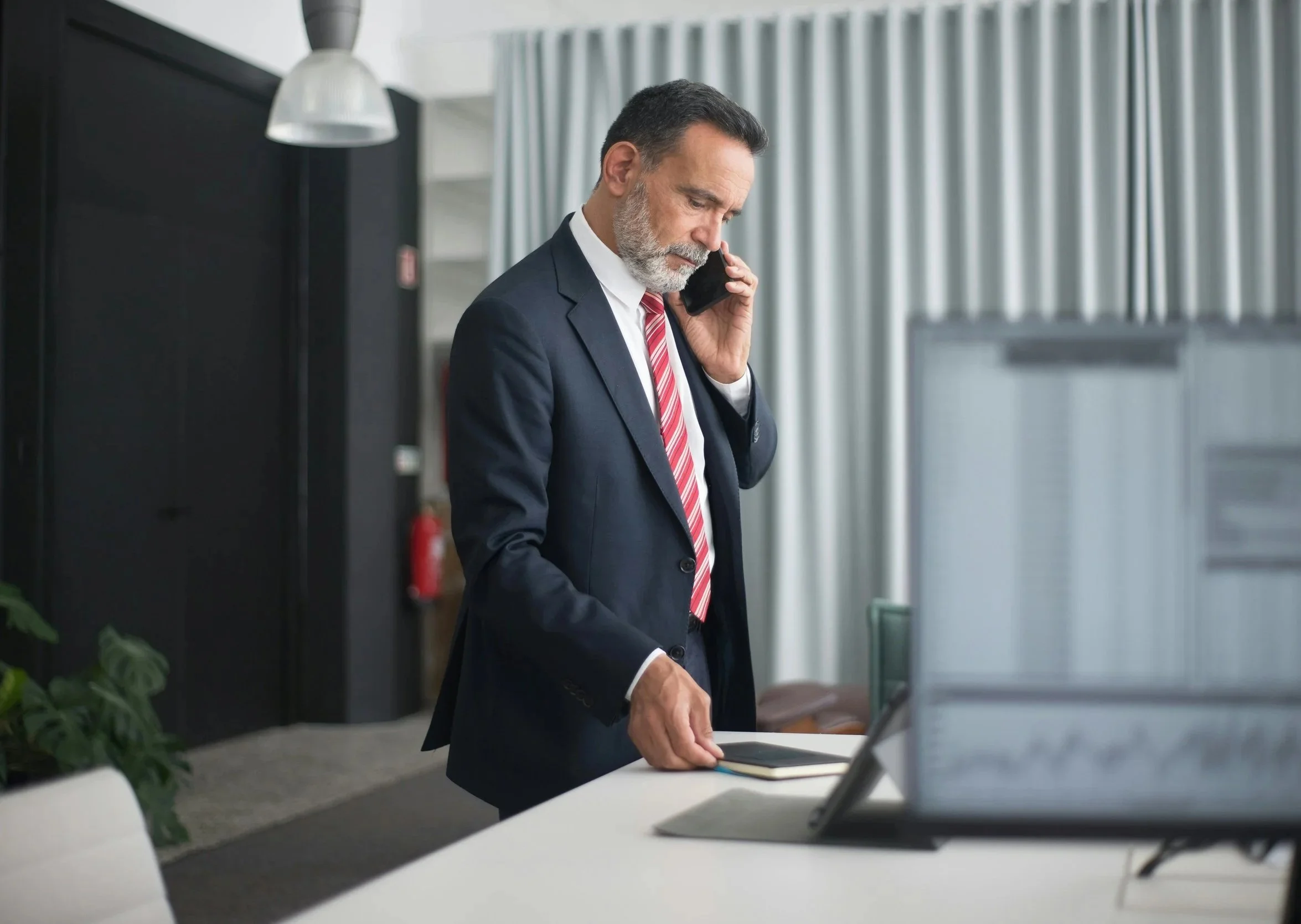 A middle-aged man in a business suit with a striped tie is standing at a desk, talking on his cellphone with a serious expression.