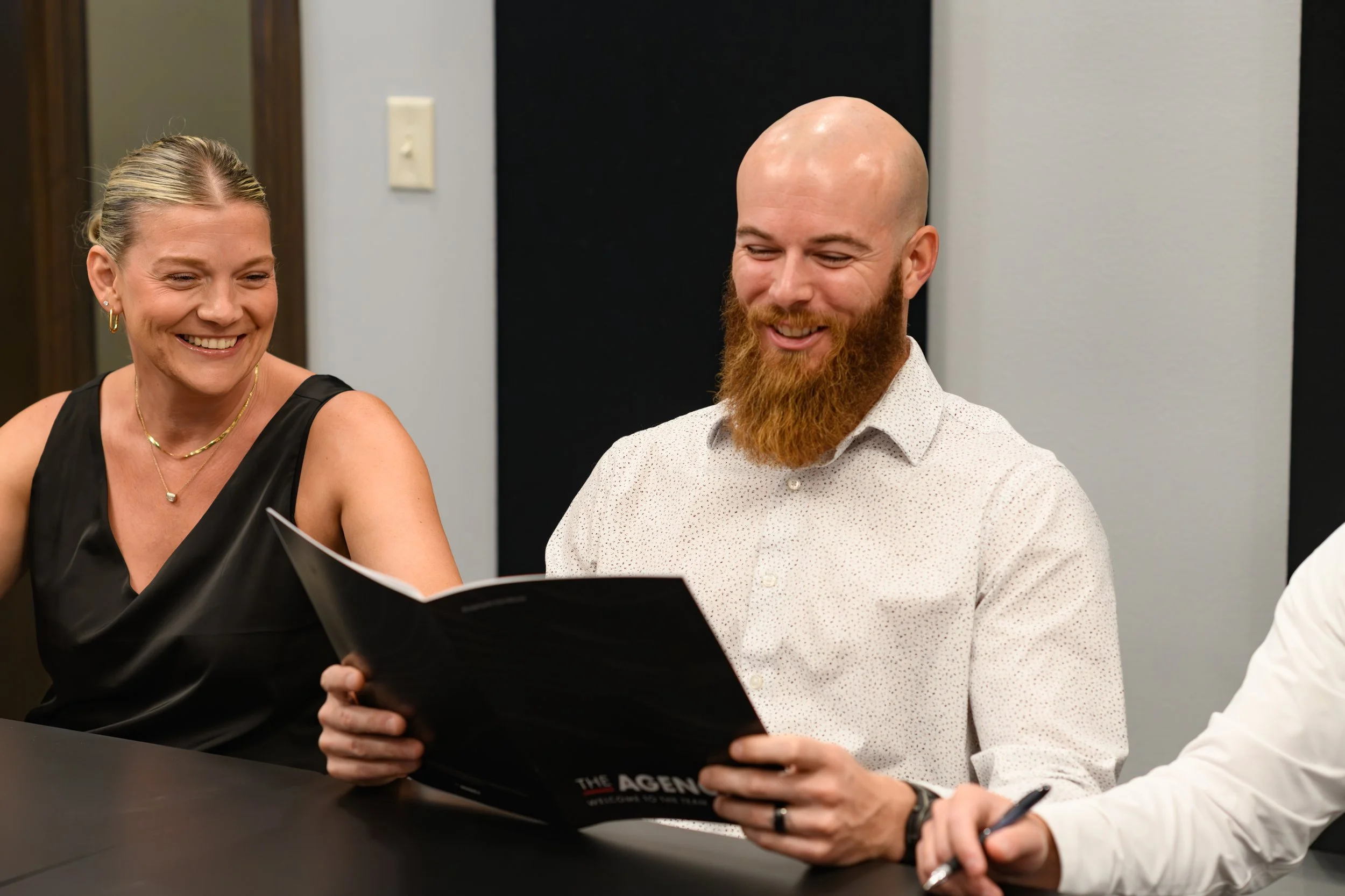Two people, a woman and a man, sitting at a table and looking at a magazine. They are smiling and appear to be having a good time.