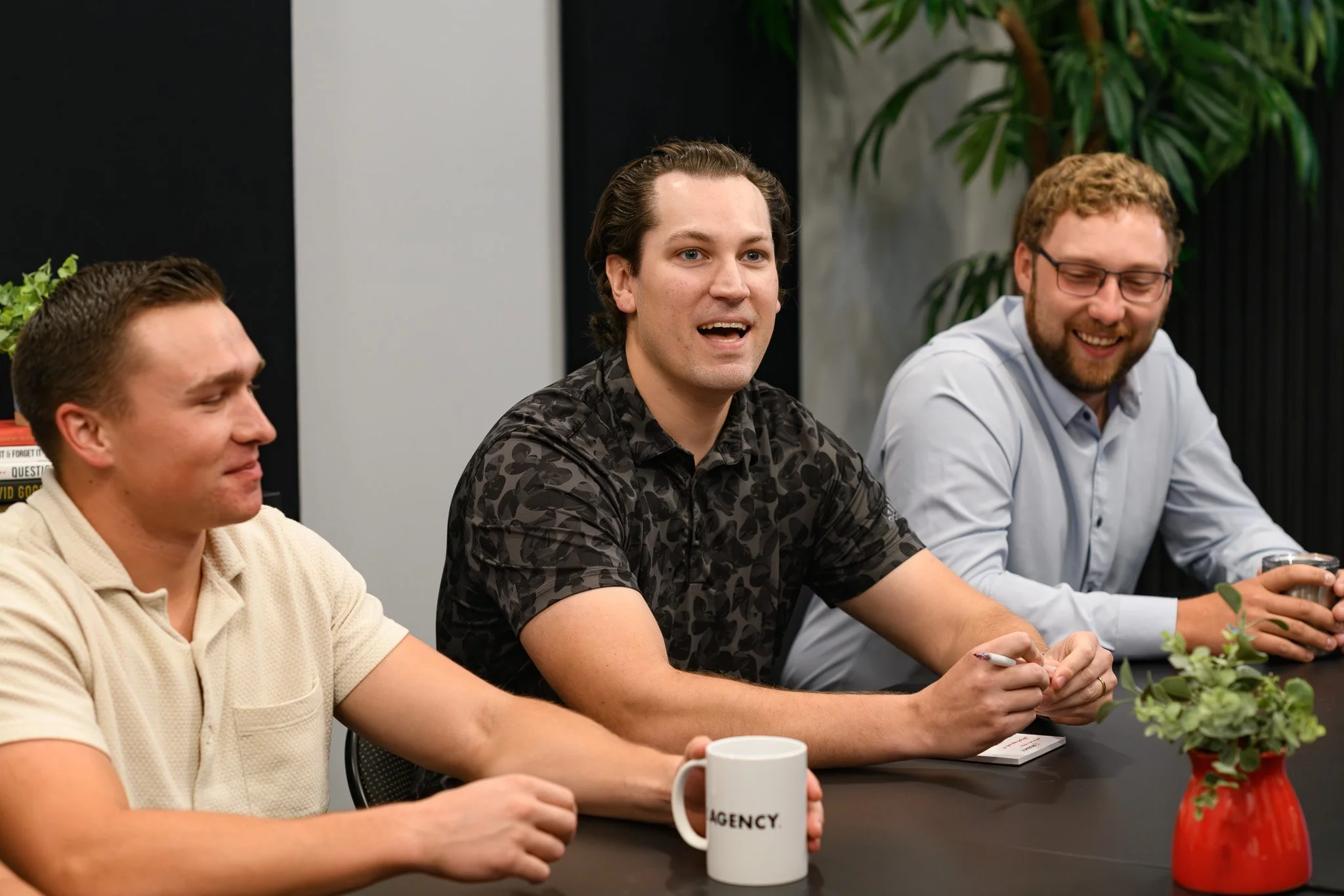 Three men sitting at a conference table in a meeting room, engaging in conversation and smiling, with plants and books visible in the background.