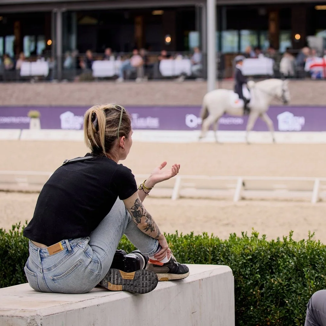 Em from Bolt&Bay Media is sitting on a bench watching a horse in an indoor arena after taking photos of the competition.