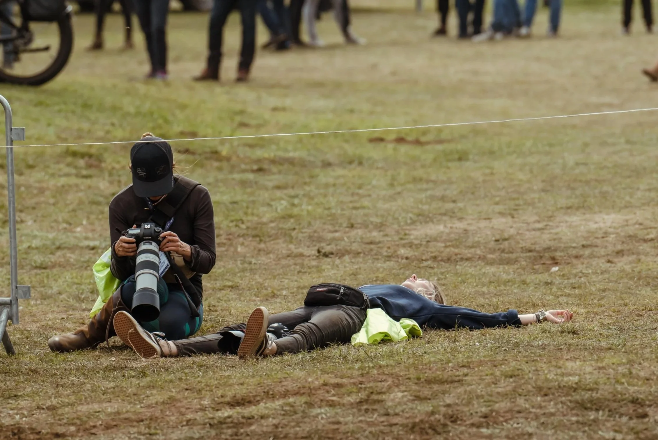 Em from Bolt&Bay Media lying on the ground at the FEI Eventing European Championships with a camera and a person kneeling next to them looking at their pictures on camera, with a group of people in the background.
