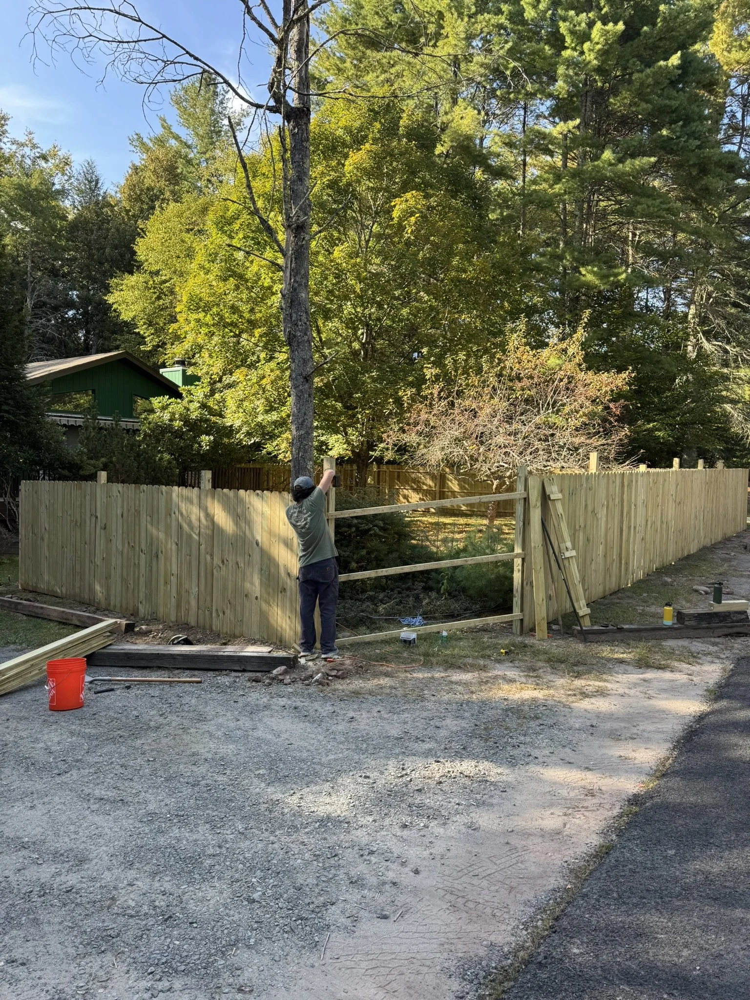 Person constructing a wooden fence beside a gravel driveway, with a green house and trees in the background.