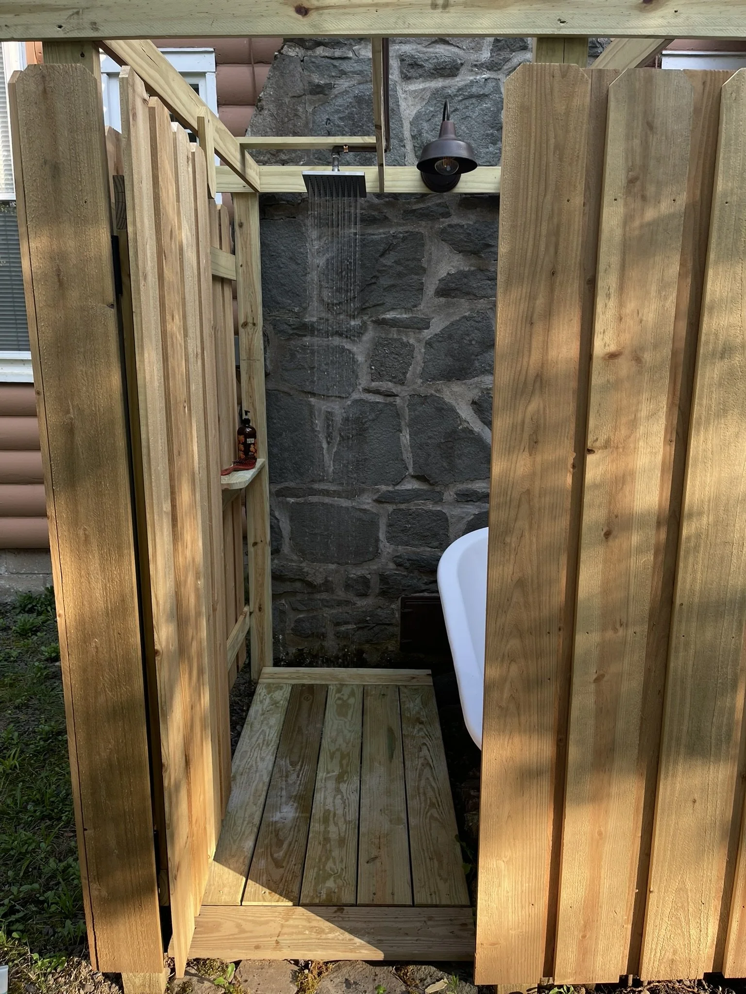 An outdoor shower with a wooden enclosure, a stone wall with a rainfall showerhead, and a small shelf with personal items, partially obscured by a white bathtub.