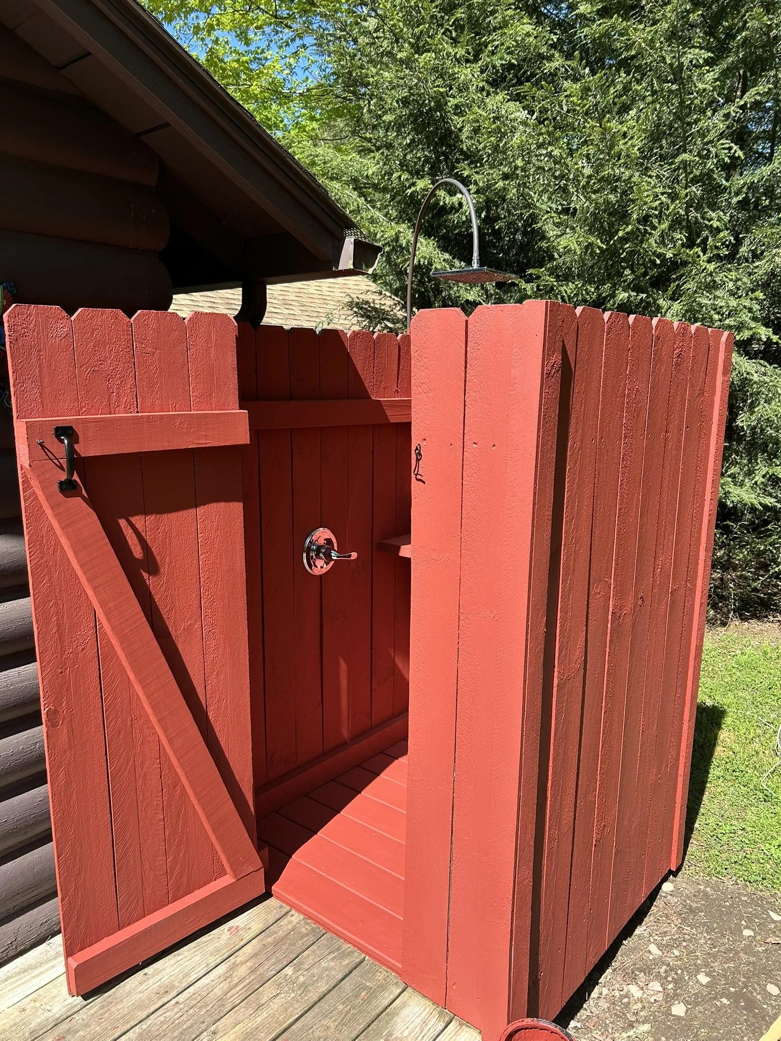 An outdoor red wooden shower with a showerhead, a door, and a small shelf inside, situated on a wooden deck with greenery in the background.