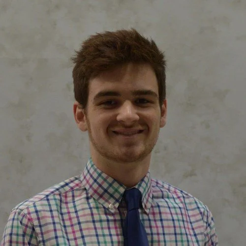 A young man with brown hair, wearing a checkered shirt and a blue tie, smiling at the camera against a plain, light-colored background.