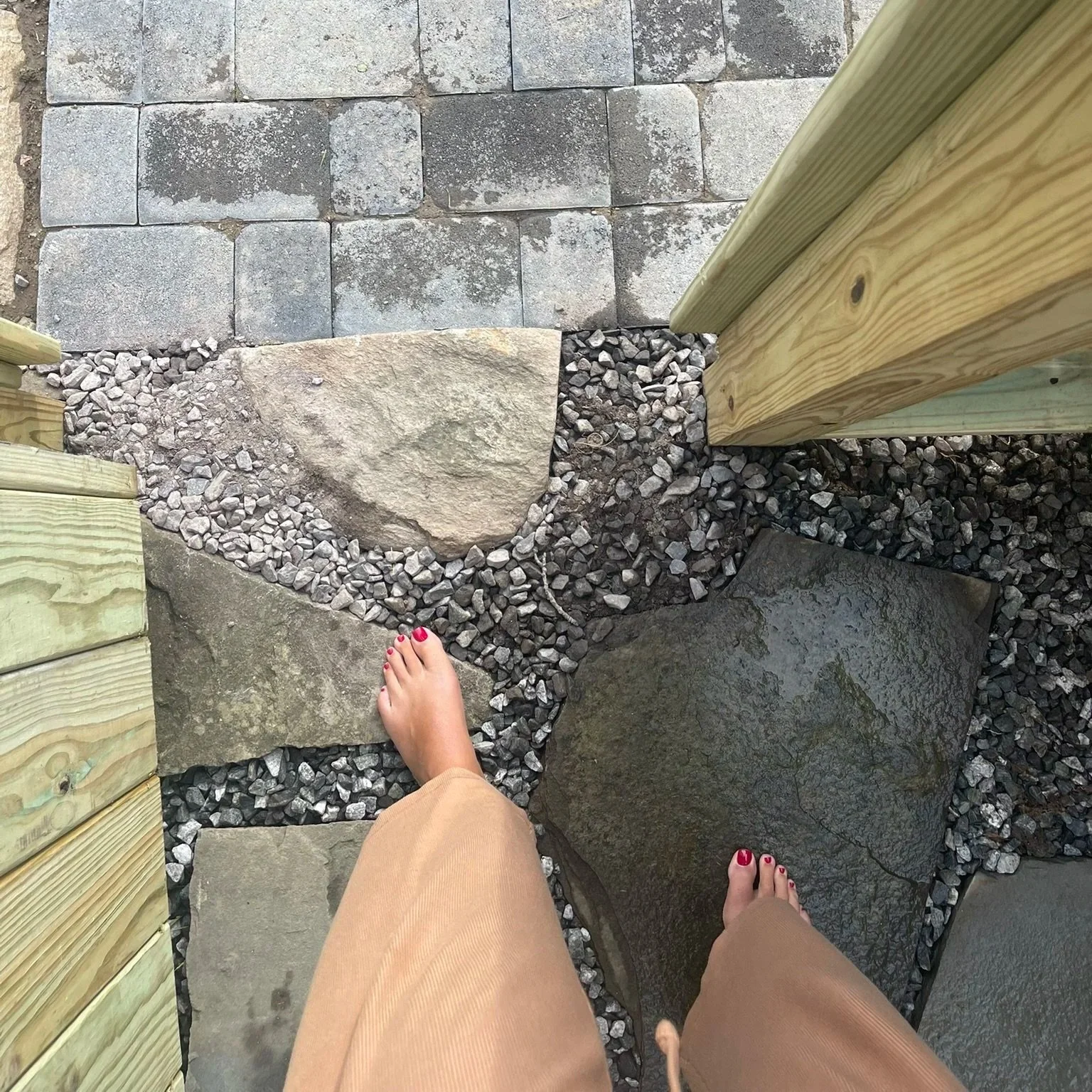Person walking on a pathway with large stones, gravel, and pavers, showing bare feet with pink-painted toenails.