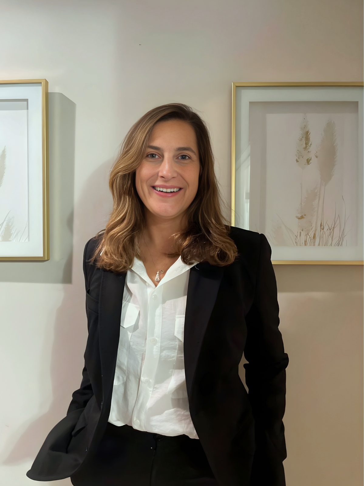 A smiling woman with shoulder-length wavy brown hair, wearing a black blazer over a white blouse, standing in front of a white wall with framed artwork featuring abstract plant designs.