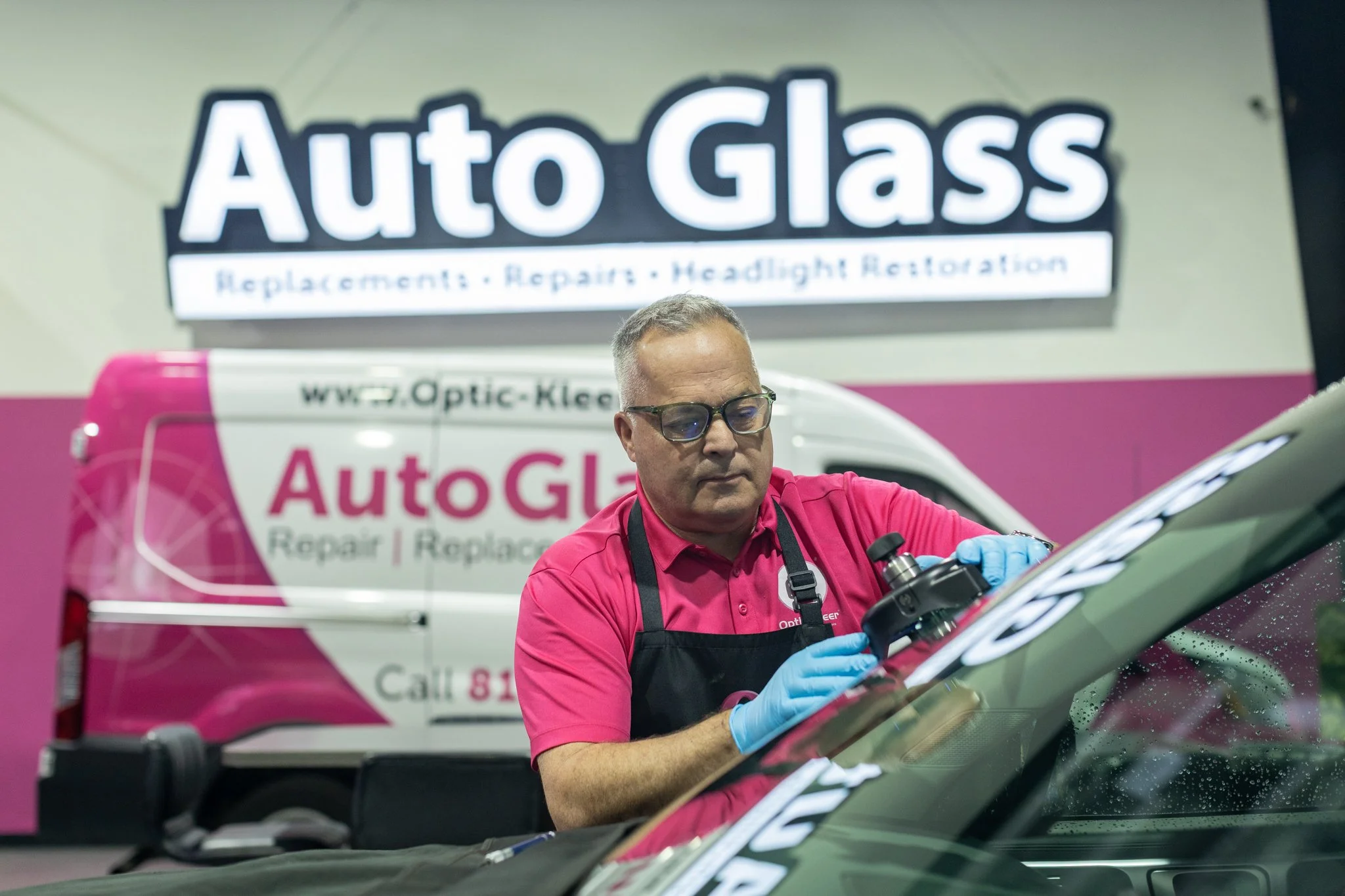 A man wearing glasses and blue gloves inspecting a car window, with a pink and white Auto Glass repair truck in the background.