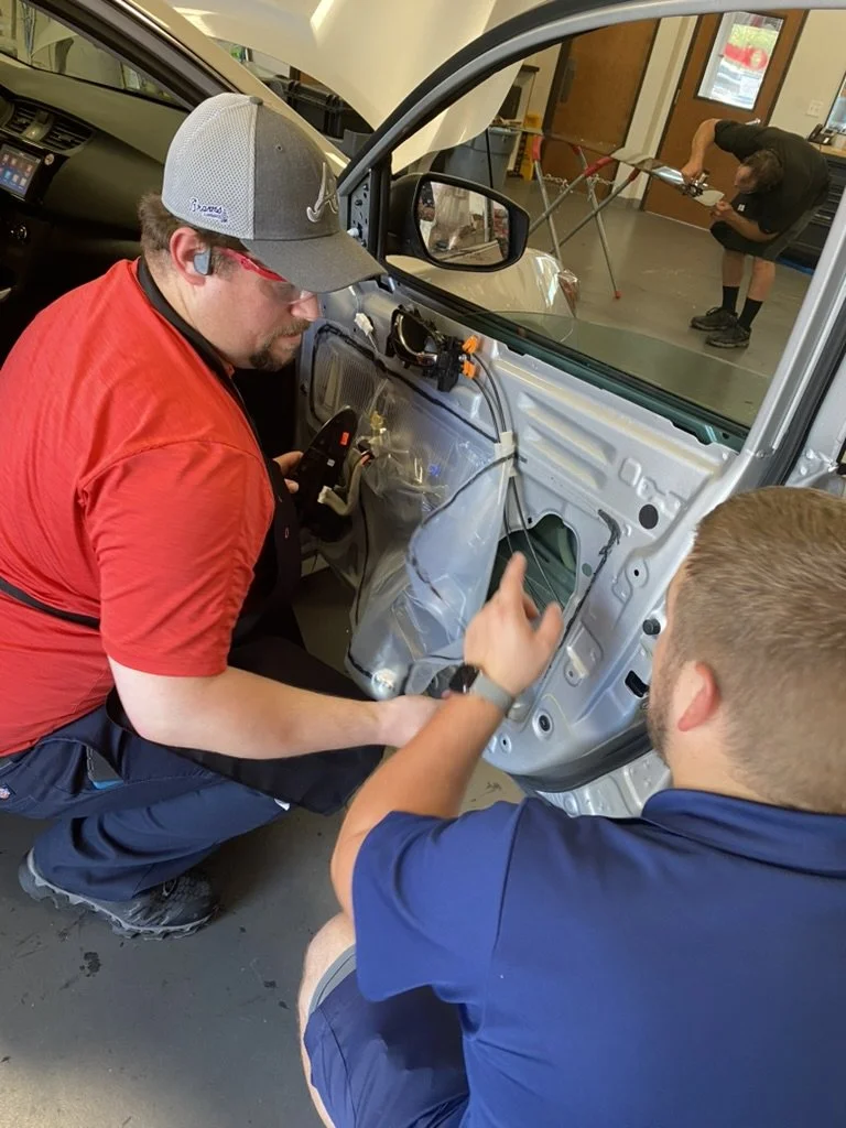 Two men working on a car door inside a garage, with one pointing at the door and the other holding a tool. In the background, another person works on a different task.