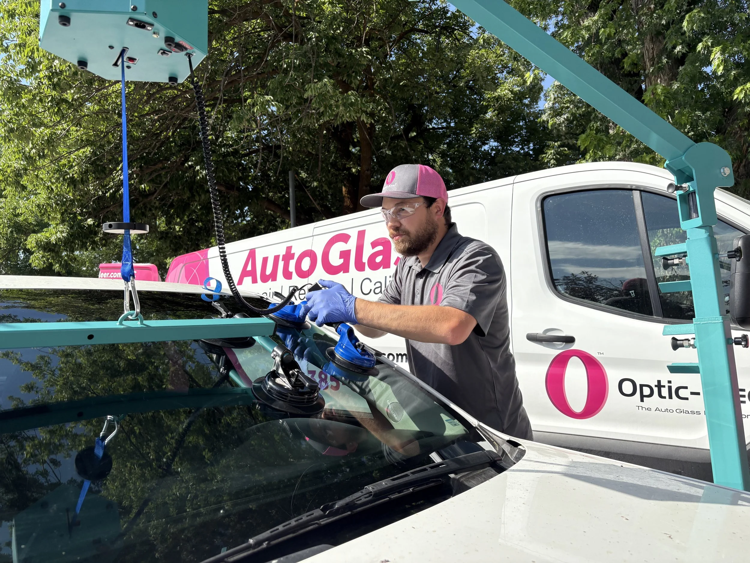 A technician repairing a car windshield using a glass installation or repair machine with an Auto Glass company truck in the background.