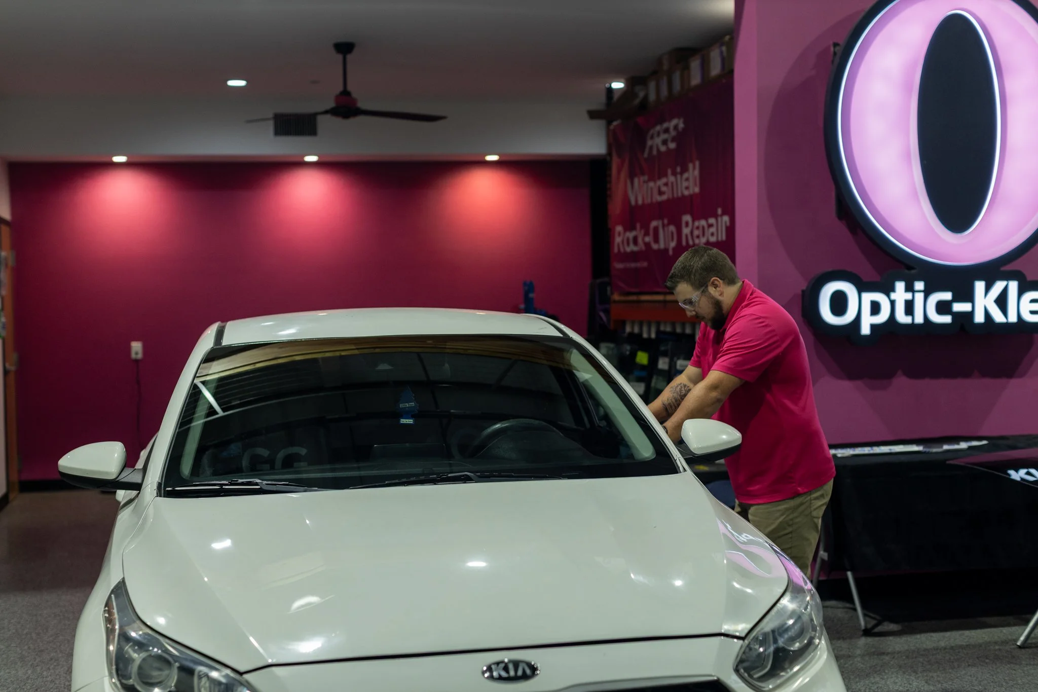 A man wearing glasses and a pink shirt is working on a white Kia car inside an indoor facility with pink walls. There is a large illuminated signage for Optic-Kleer on the right side of the image.