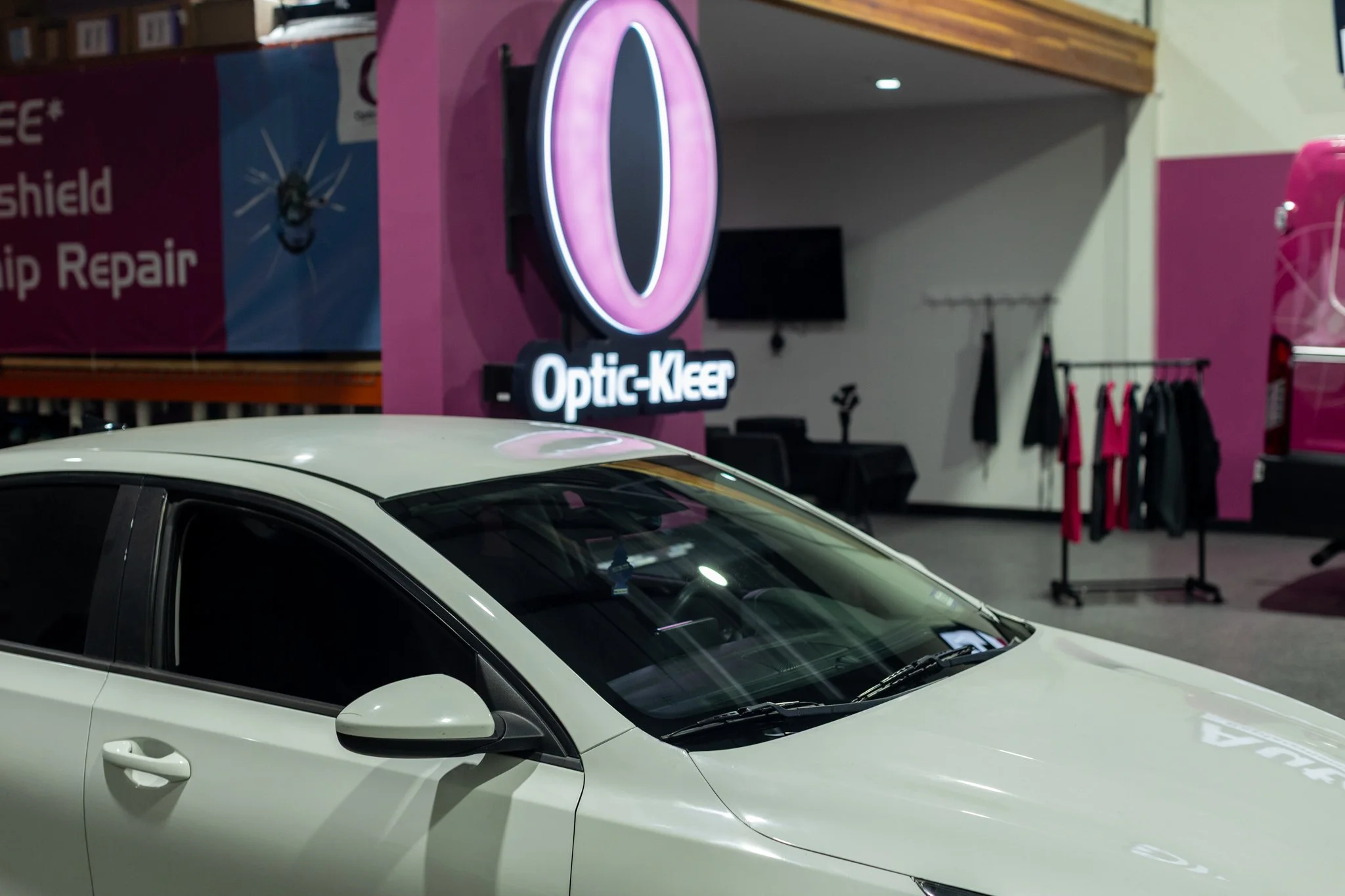 A white car parked indoors near a pink wall with a neon sign reading 'Optic-Kleer'. In the background, there are clothes hanging on racks and a TV mounted on the wall.