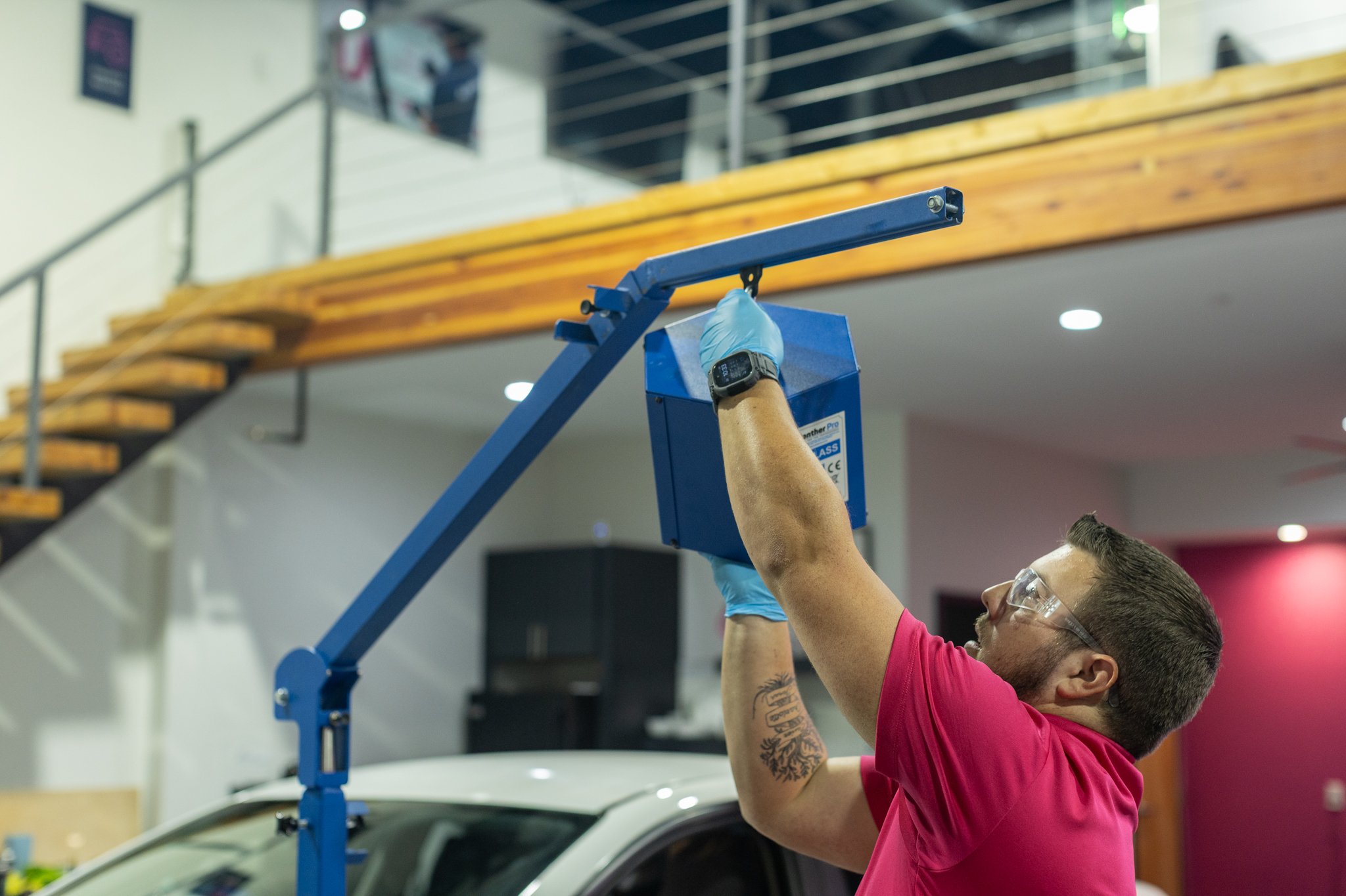 A man wearing safety goggles, blue gloves, and a red shirt is installing or repairing a blue device with a long arm in an indoor space with a staircase and a car in the background.