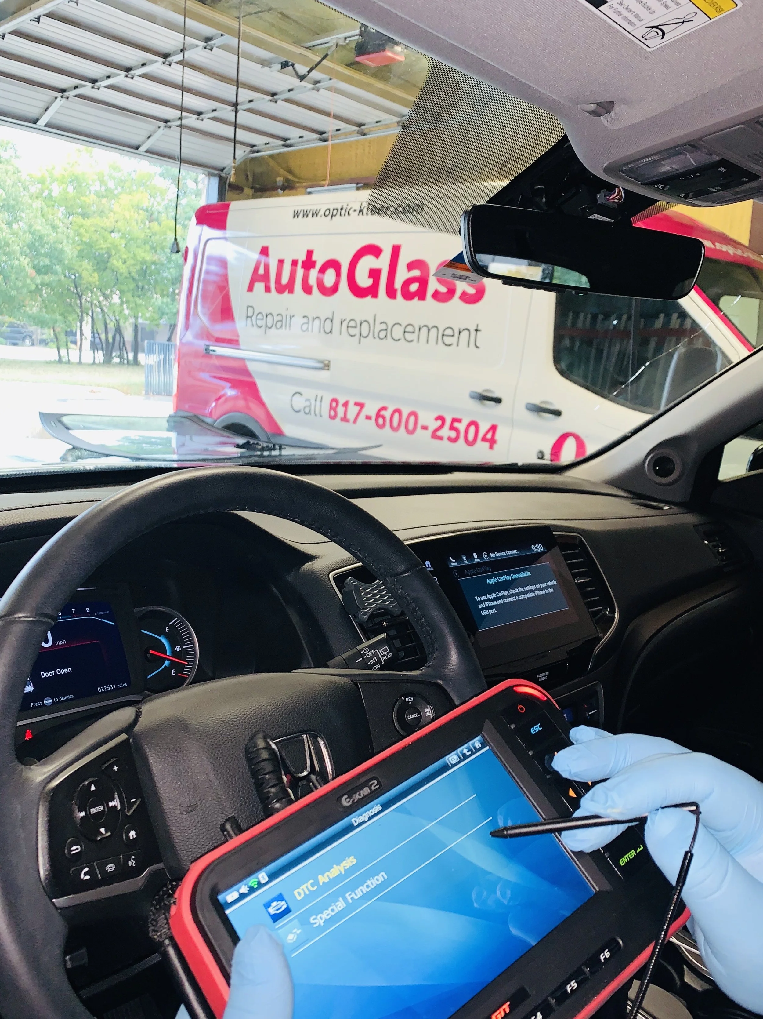 Inside a vehicle, a mechanic is diagnosing a car issue with a handheld scanner. The vehicle's dashboard shows an open door notification. Outside the vehicle, a white van with pink and black lettering advertises AutoGlass repair and replacement, with a contact phone number.
