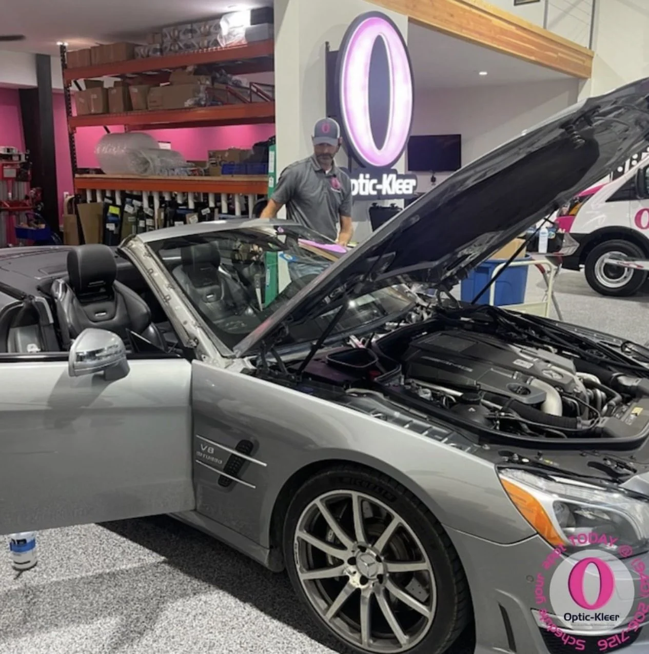 A silver Mercedes-Benz convertible with its hood open and a V8 engine visible. A man in a gray cap and gray shirt is working on the car inside an automotive shop with pink and black signage that says "Optic-Kleer." The shop has shelves with boxes and supplies in the background, and a promotional sign with a large pink "O" and the words 'Schedule Your Service Today'.
