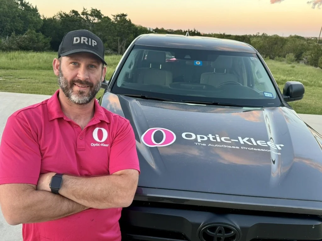 A man in a pink polo shirt and a black cap with the word 'DRIP' stands with arms crossed next to a gray vehicle marked with 'Optic-Kleer' and a large pink and black eye logo, outdoors during sunset.