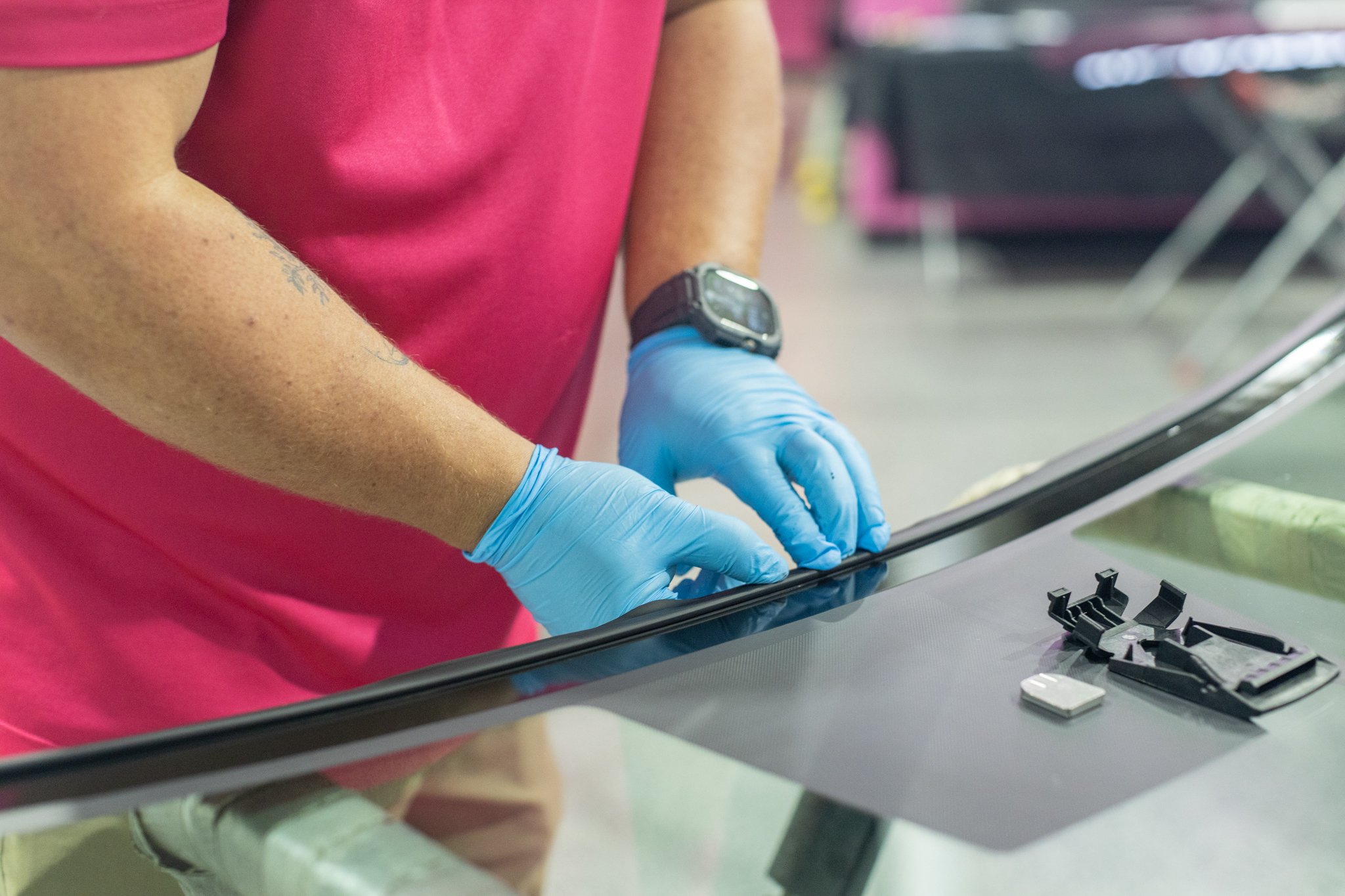 Person wearing a pink shirt and blue gloves working on a car window, with small tools and parts on the work surface.
