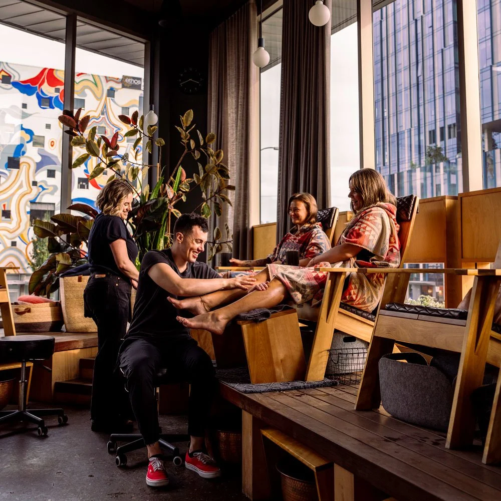Two people in colorful robes sit in chairs and have their feet massaged by two people dressed in black.