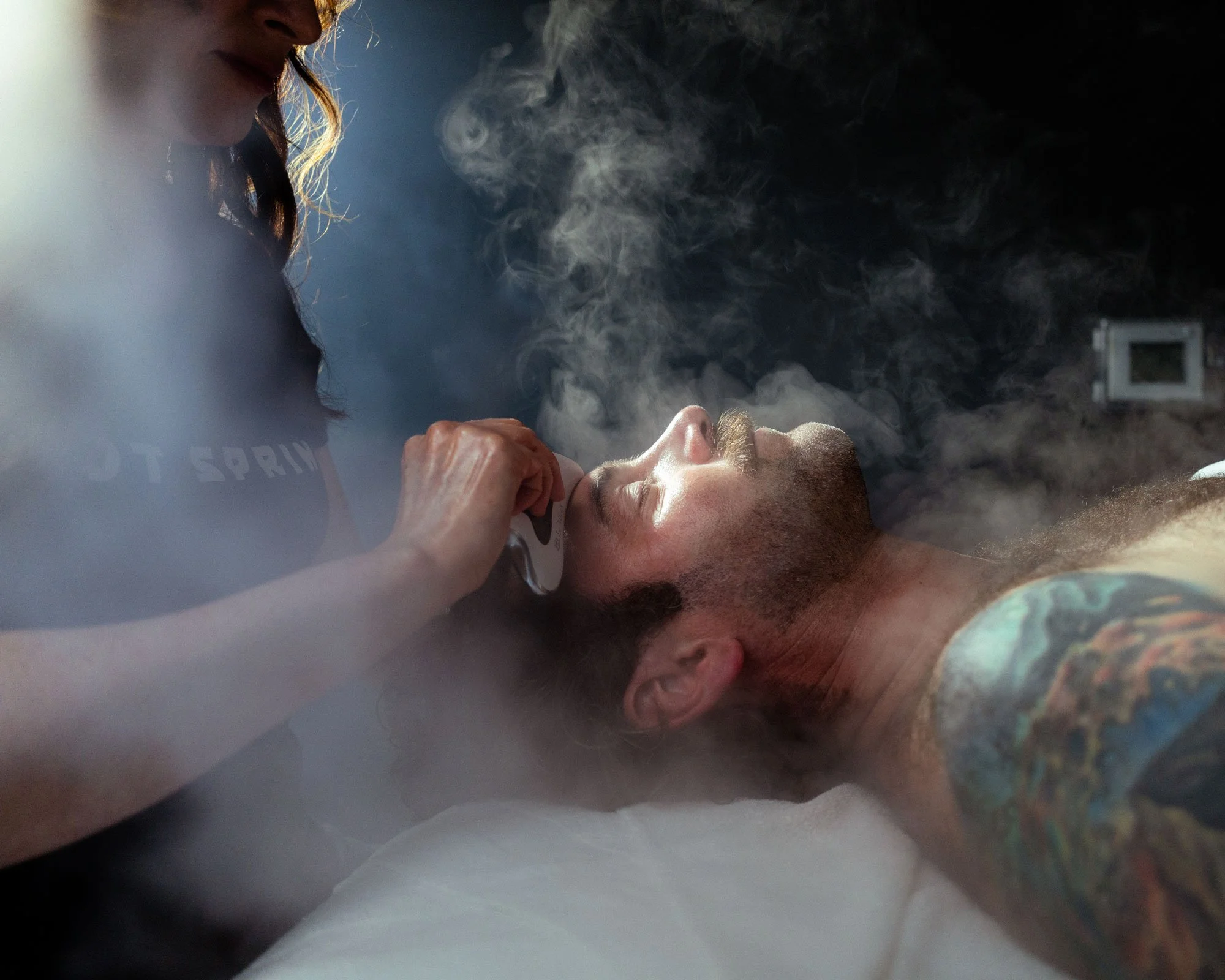 A man lies on a table with steam around his face, while an esthetician massages his face, receiving a Detoxifying Facial at Knot Springs