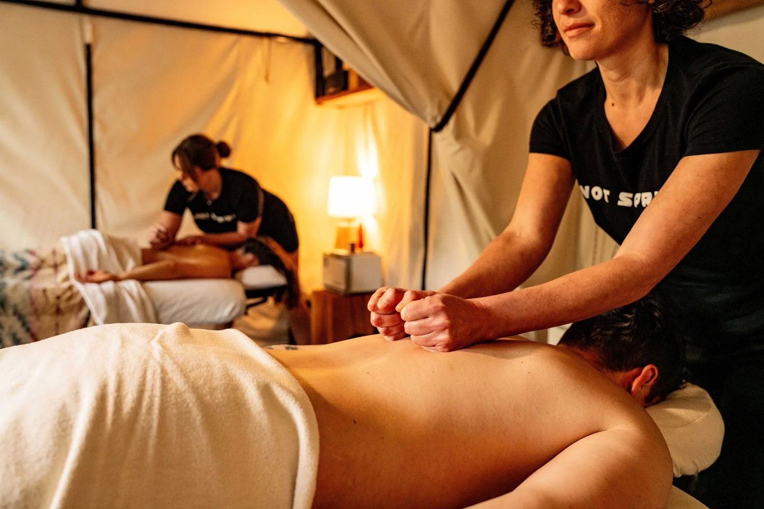 Two people lie face down on massages tables and receive massages on their backs from massage therapists
