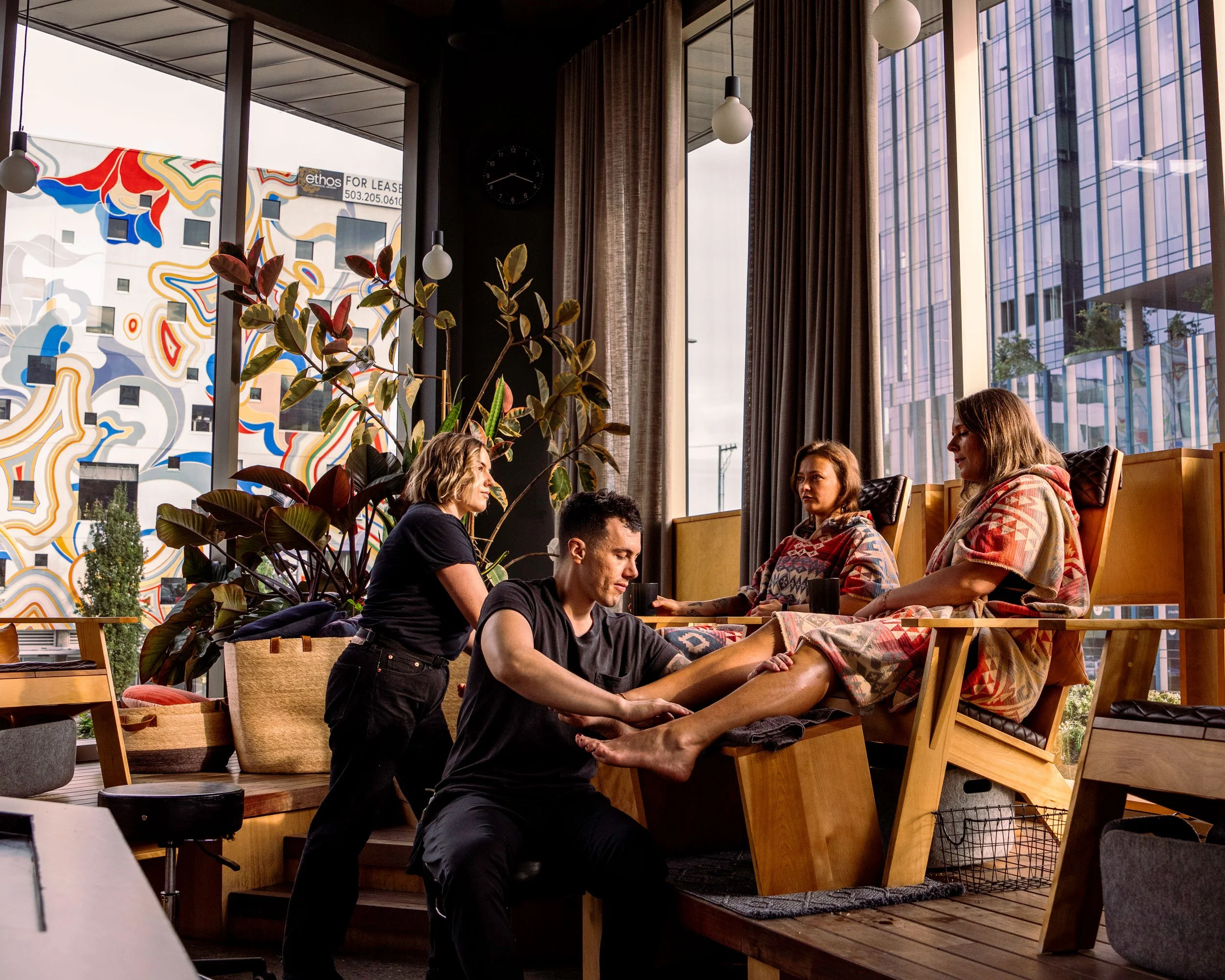 Two women in brightly colored robs sit in wooden chairs and receive foot massages from two massage therapists
