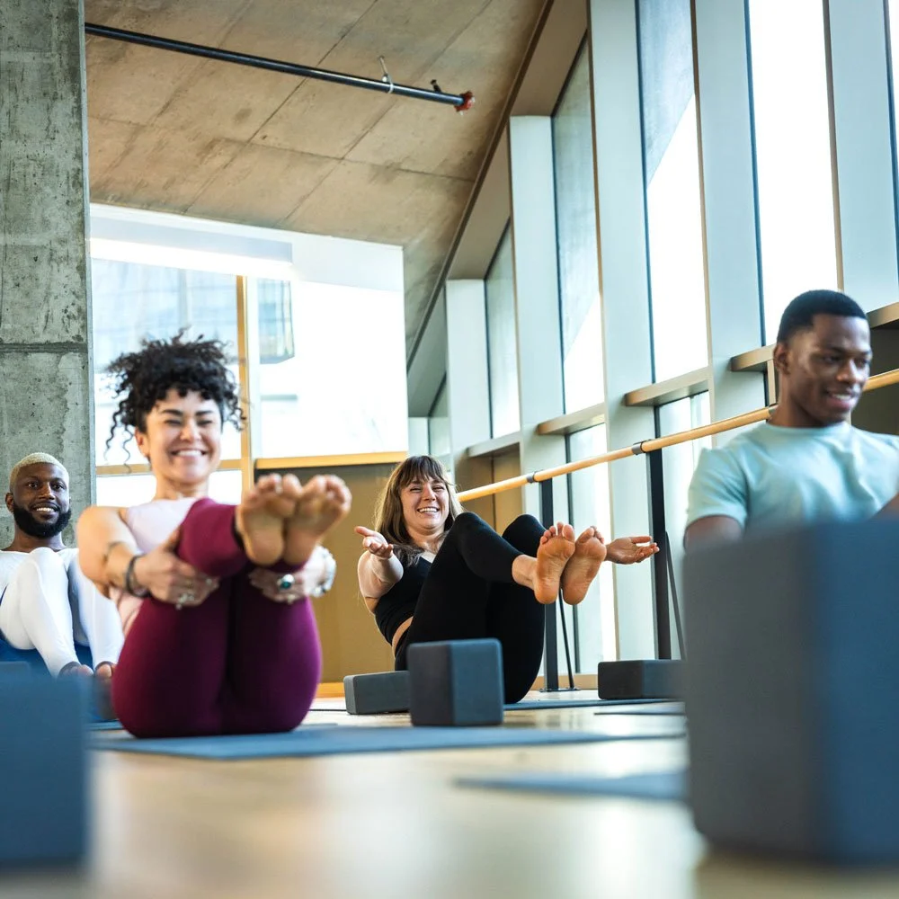 Four people sit on the ground in a sunny yoga studio with their arms and legs raised in a stretch