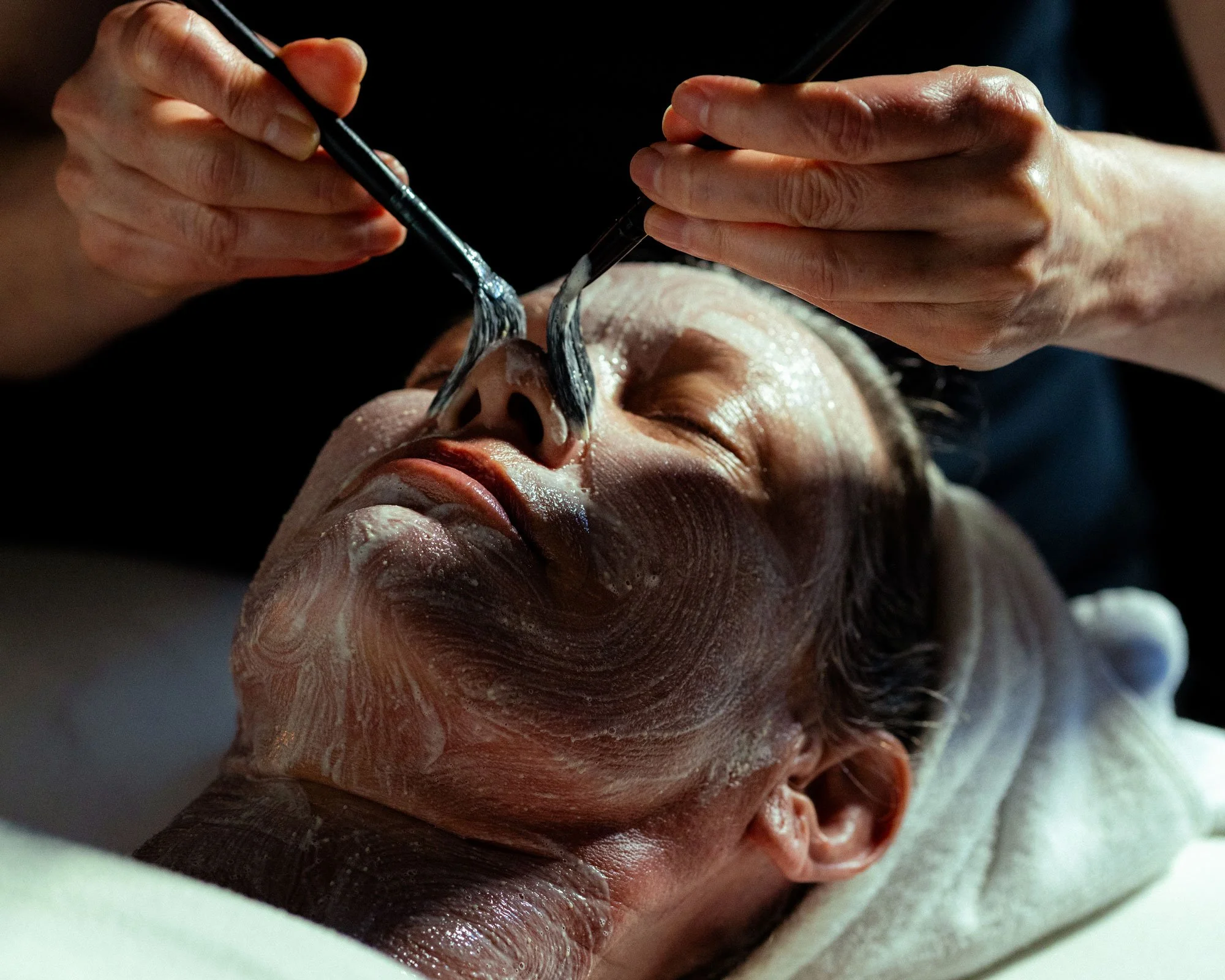 A woman lies on a treatment table and has light colored serums applied to her nose with two brushes, receiving a Brightening Facial treatment at Knot Springs