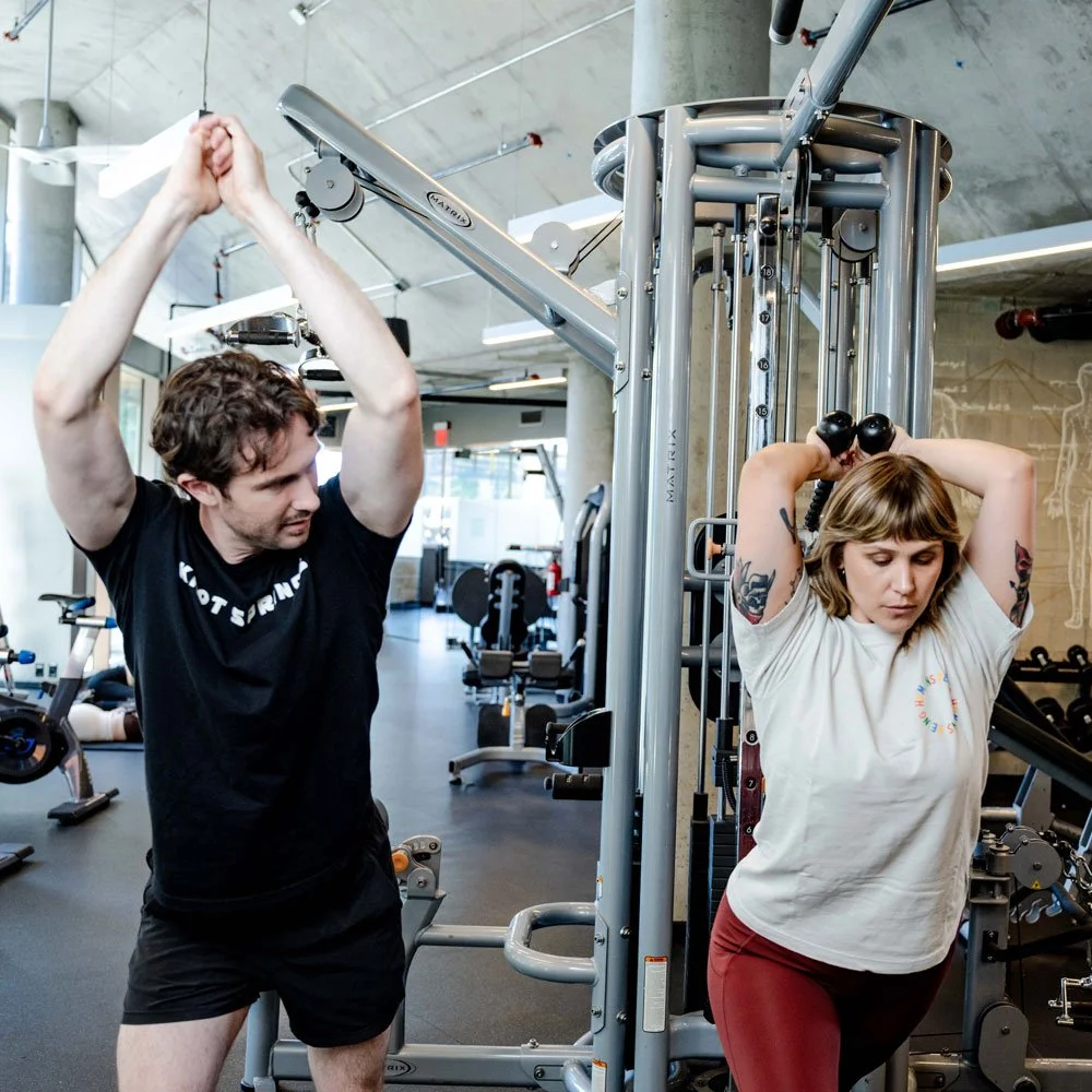 A trainer shows a client how to lift weights overhead in a gym