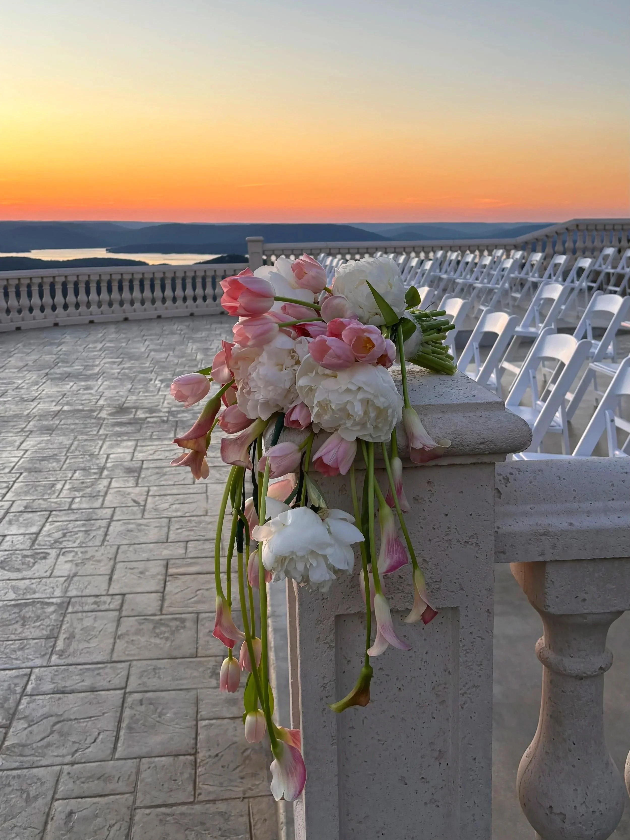 A bouquet of pink and white flowers laid on a stone railing with white chairs in the background, overlooking a sunset over a body of water and hilly landscape.