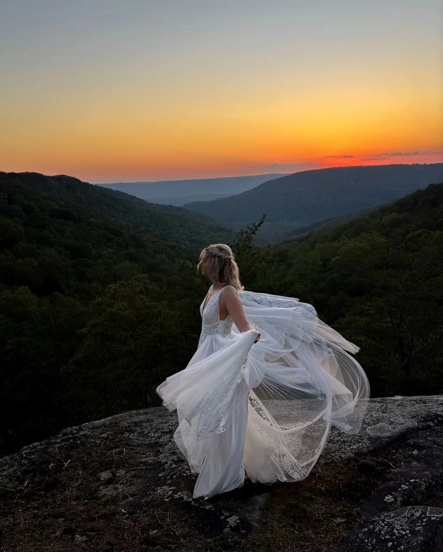 A Bride in a white wedding dress standing on a rock overlooking a mountain landscape at sunset with a colorful sky on her wedding day, captured by her wedding content creator.