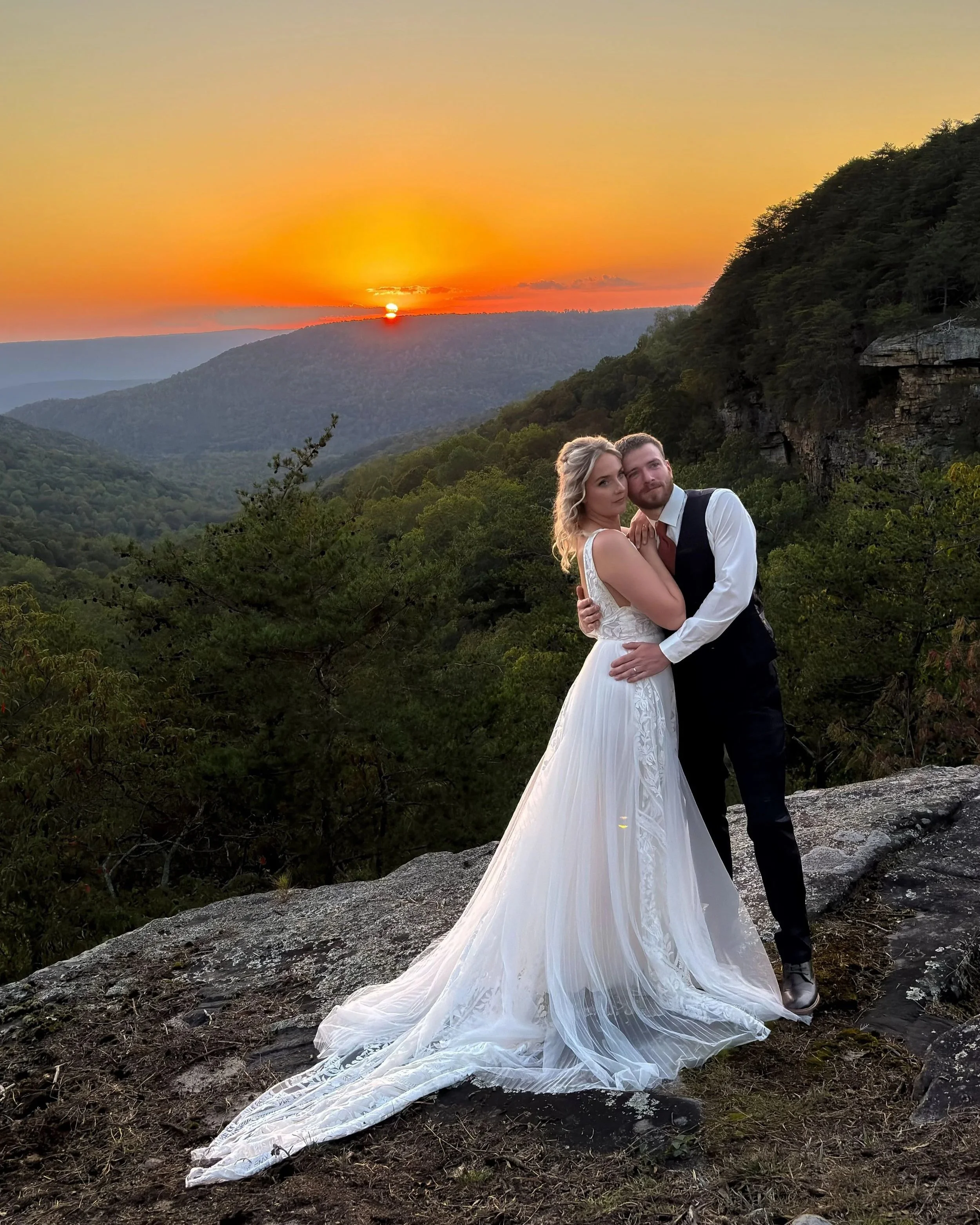 A newlywed couple stands close together on a rocky ledge overlooking a lush green valley with a sunset in the background. The bride is in a flowing white wedding gown, and the groom is in a black vest and white shirt. The sunset features an orange an
