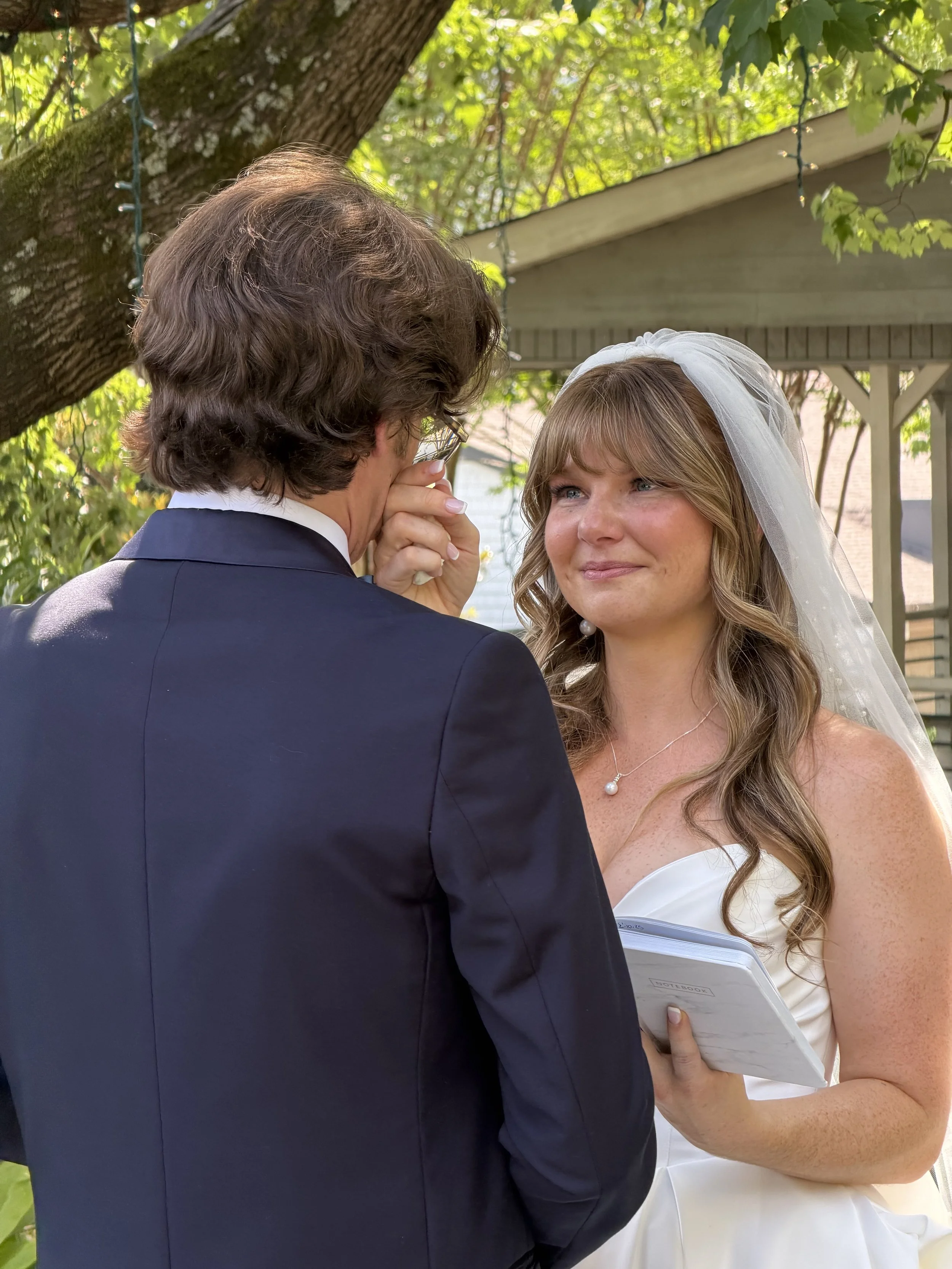 A bride and groom during their wedding ceremony outdoors, with the groom whispering to the bride under a leafy tree.
