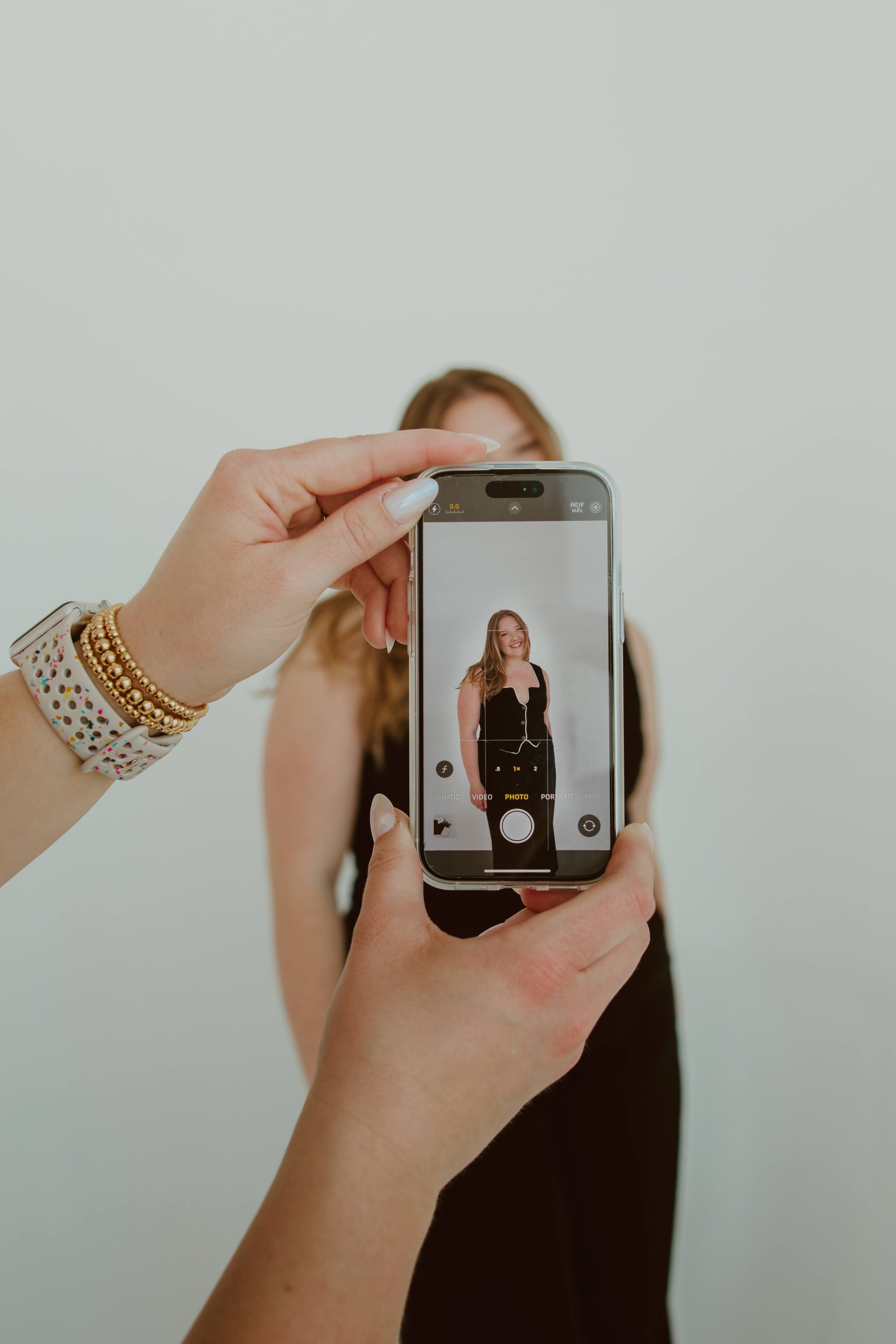 Close-up of a woman's hand holding a smartphone with a woman in a black dress on the screen, taking a photo against a plain white background.