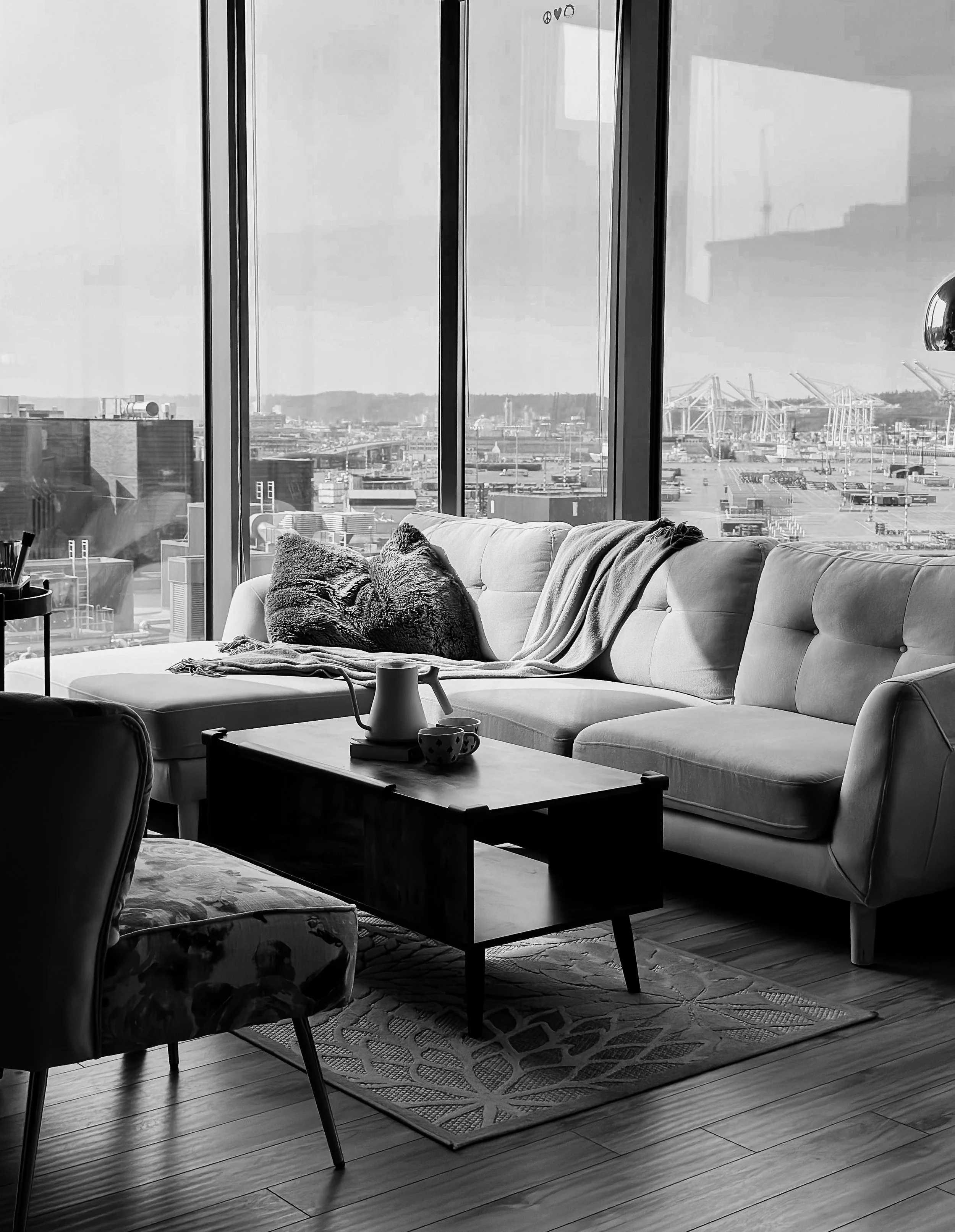 A black and white photo of a modern living room with a large window showing an airport or port with cranes and containers outside. Inside, there is a spacious sofa with pillows and a blanket, a small coffee table with a teapot and cups, and a pattern