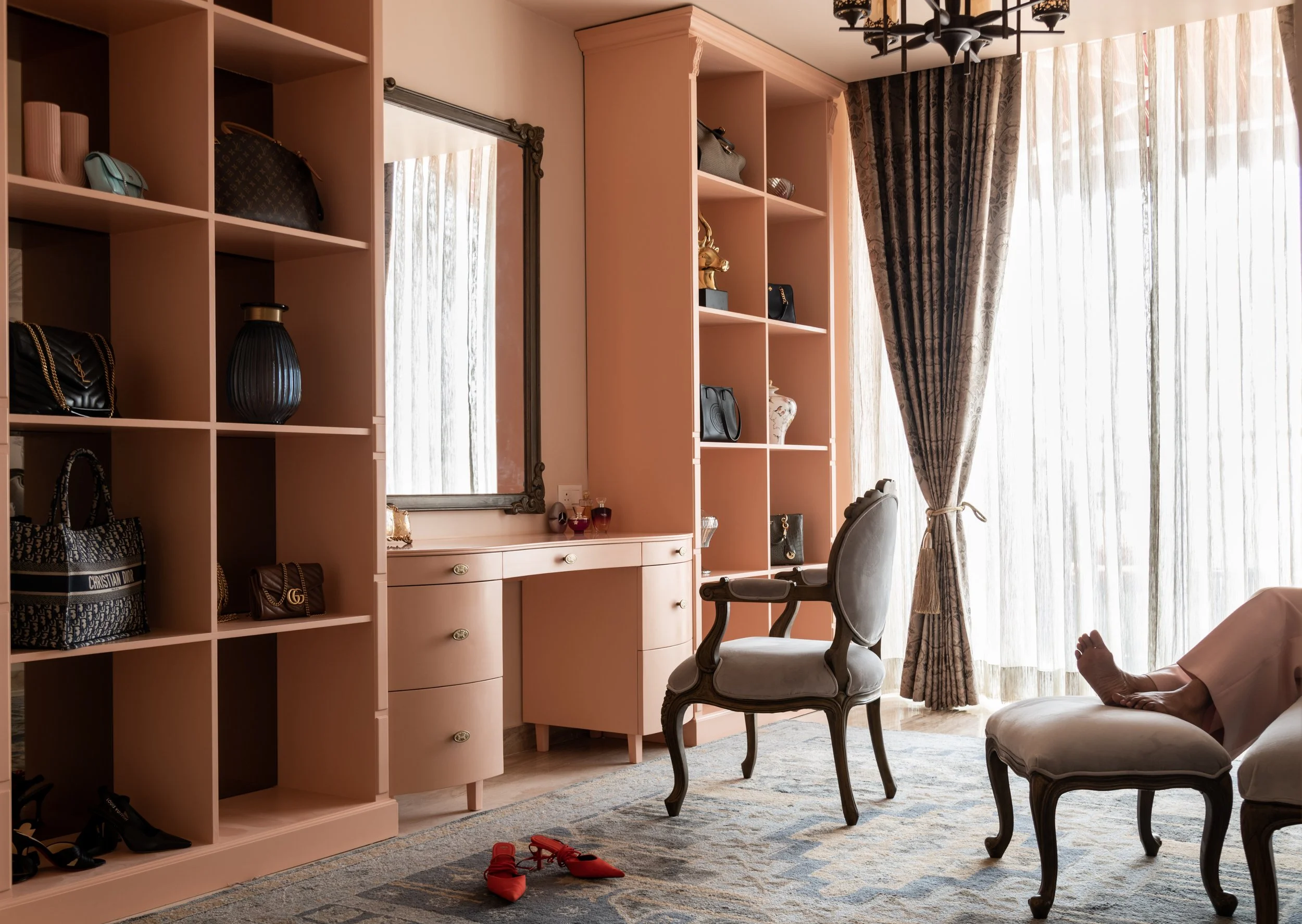 A luxurious bedroom with pink shelving units displaying handbags and decorative items, a vintage vanity with a mirror, an elegant armchair, a pair of red shoes on a patterned rug, and a large window with sheer curtains and dark drapes, with a person'