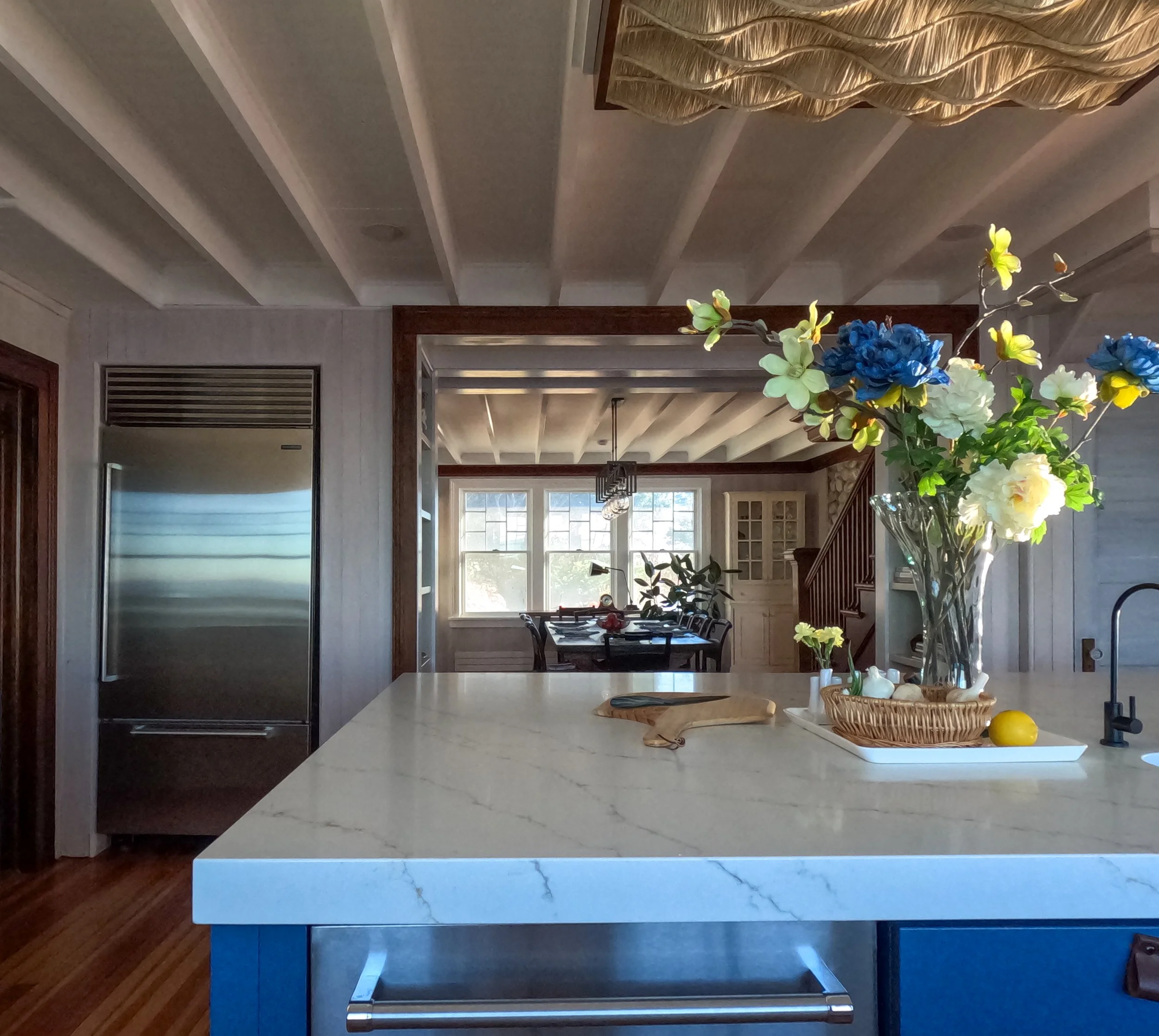 Modern kitchen with a marble island counter, a vase of colorful flowers, and a view into a dining area with large windows and wooden staircase.