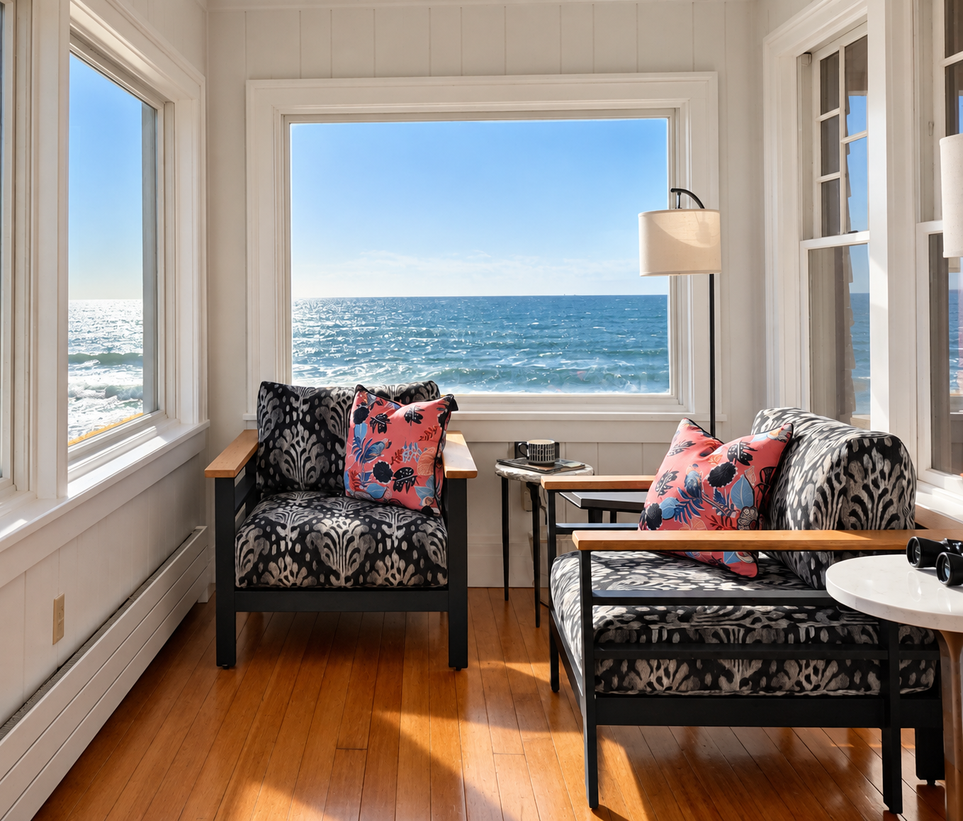 Living room with ocean view through large windows, black and white patterned armchair and sofa with floral cushions, small side tables, a floor lamp, and a hardwood floor.